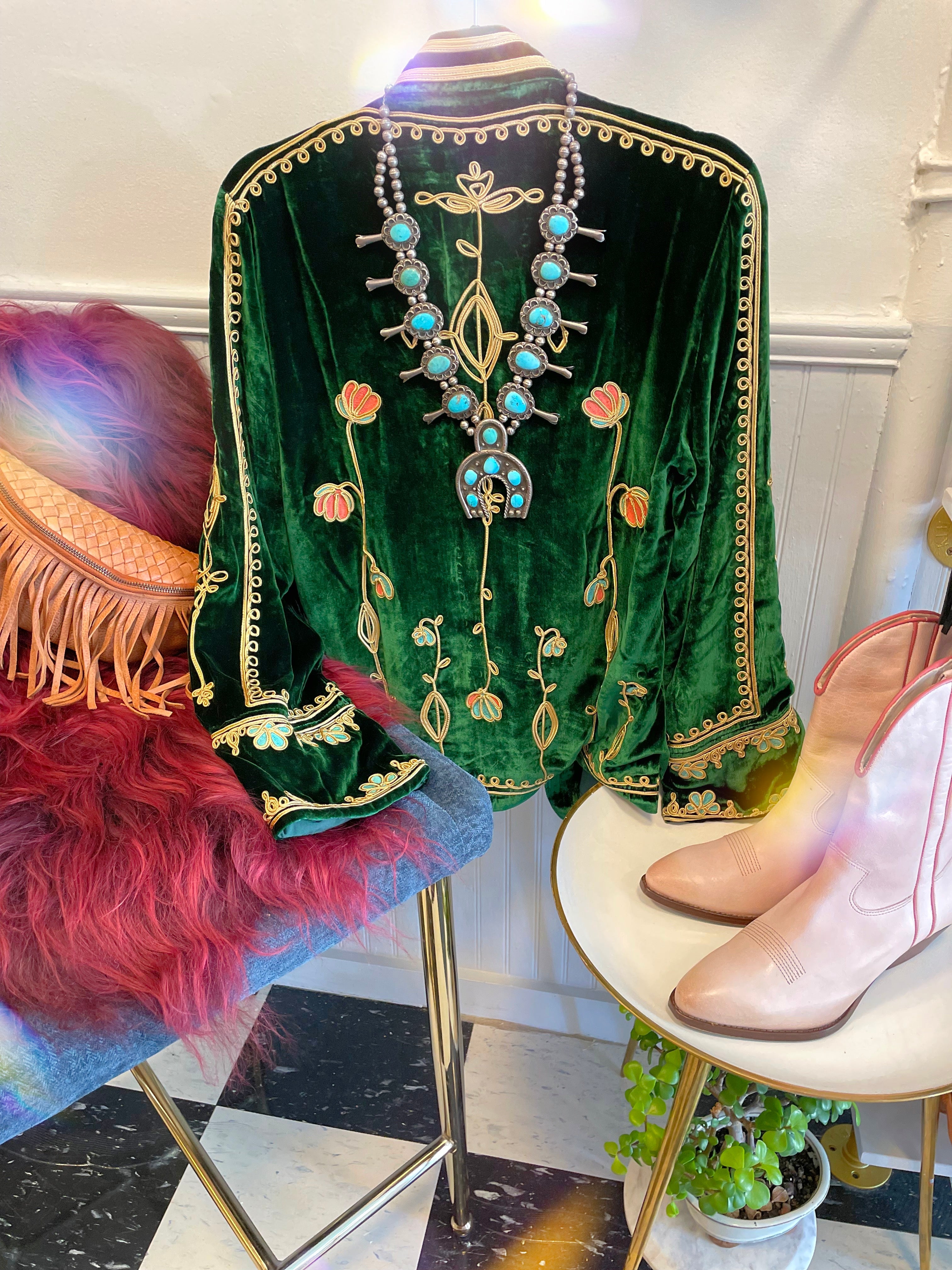 A green velvet jacket with floral embroidery, a turquoise and silver necklace, pink cowboy boots, and a fringed hat on a furry red stool, displayed on a black-and-white tiled floor.
