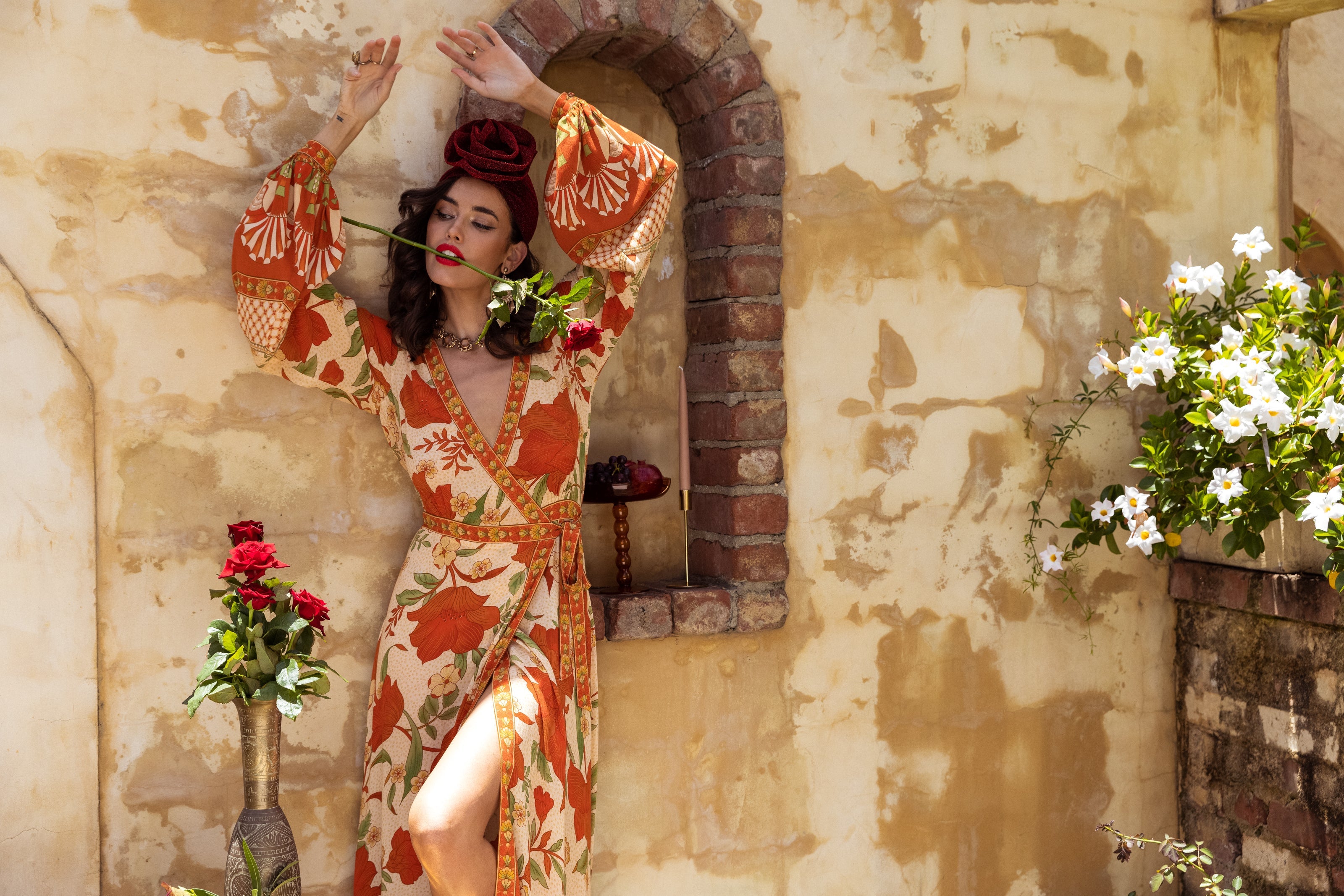 A woman in a floral dress poses against a rustic wall, holding a rose in her mouth. She has a red rose crown and stands near a vase and white flowers, evoking a romantic, vintage atmosphere.