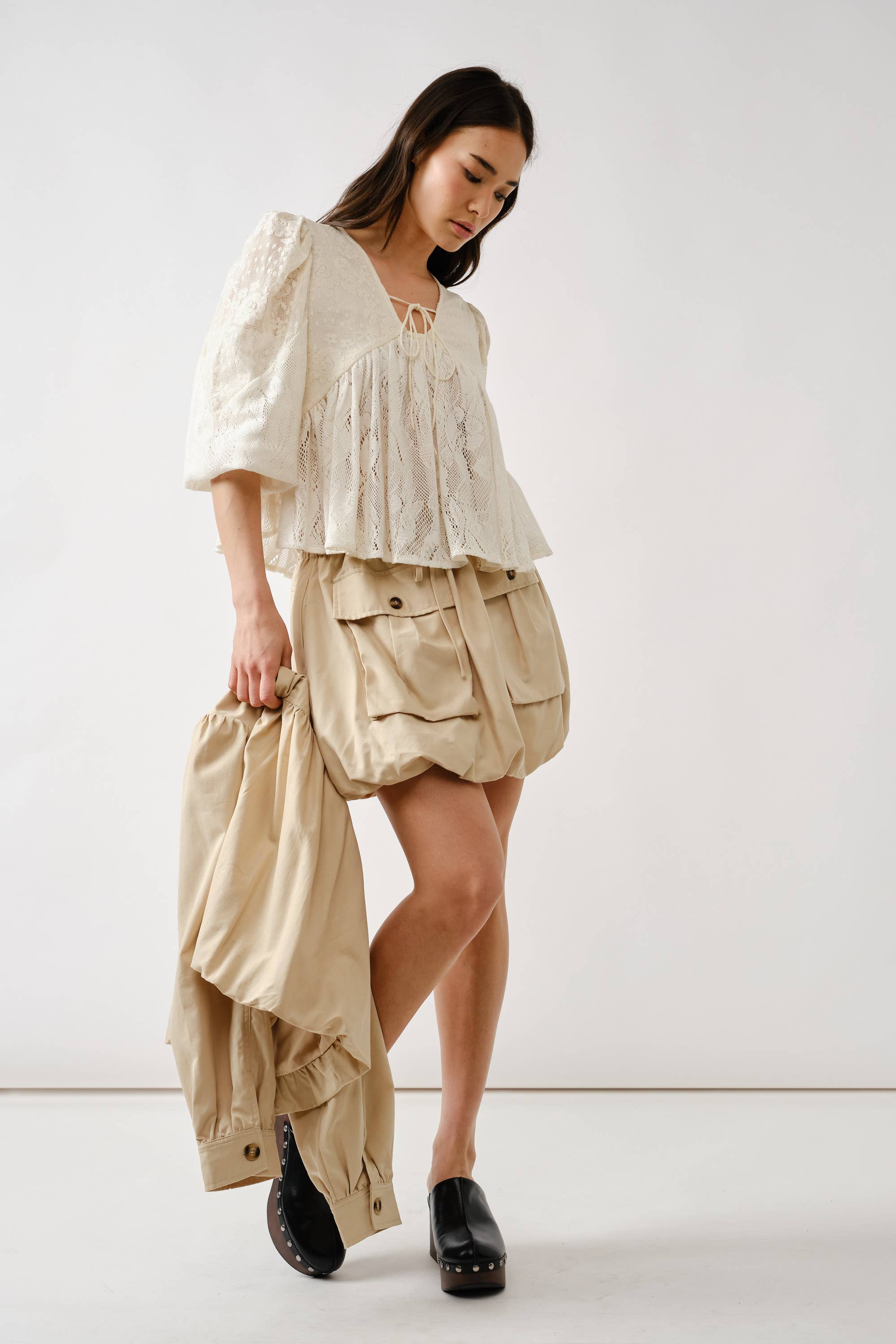 A woman wears the Aaron & Amber Long Sleeve Lace Babydoll, a loose white floral top, paired with a beige bubble skirt and black platform shoes. She holds a beige jacket and looks down against a plain white background.