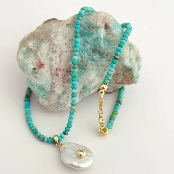 The Mar Turquezza Necklace by Tramps+Thieves, featuring turquoise beads, baroque pearls, a gemstone pendant, and a gold-filled clasp, rests atop a rough stone with blue-green hues.