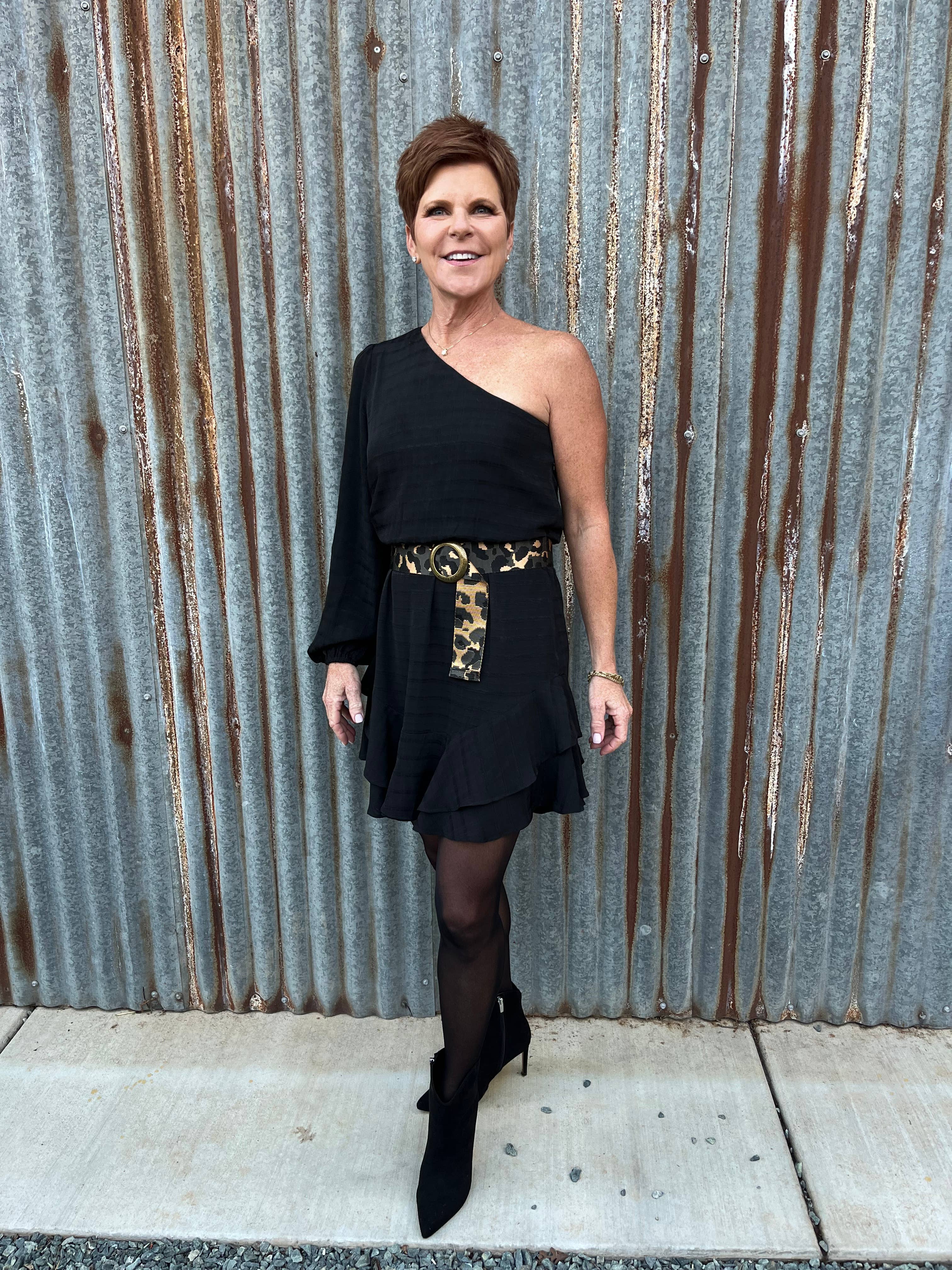 A woman with short brown hair smiles in front of a corrugated metal wall, wearing a black one-shoulder dress, sheer black tights, ankle boots, and the Ginger Blue Wild Child Leopard belt.