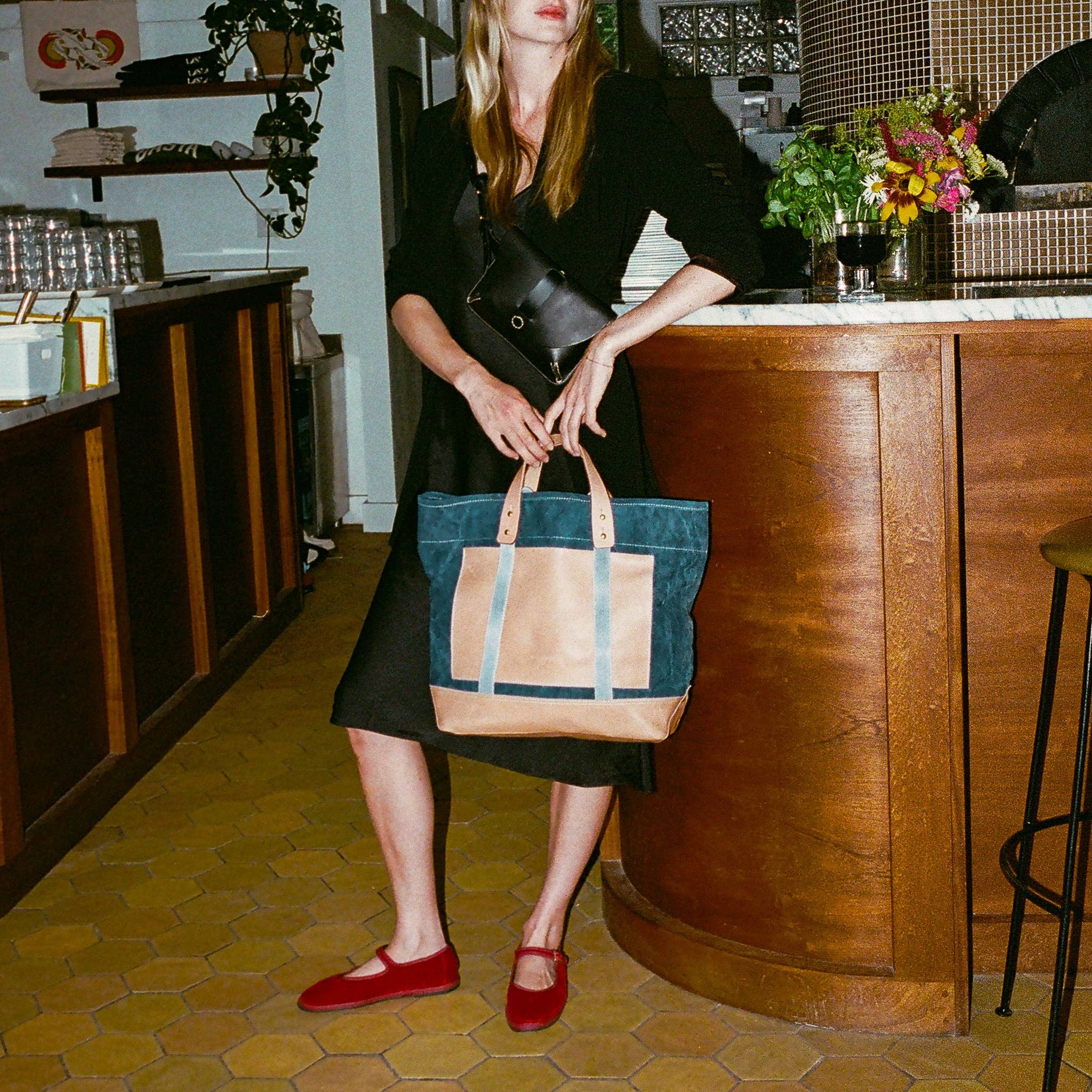 Woman in black dress holding Editor Tote | Indigo + Tan with leather handles while standing in cafe interior