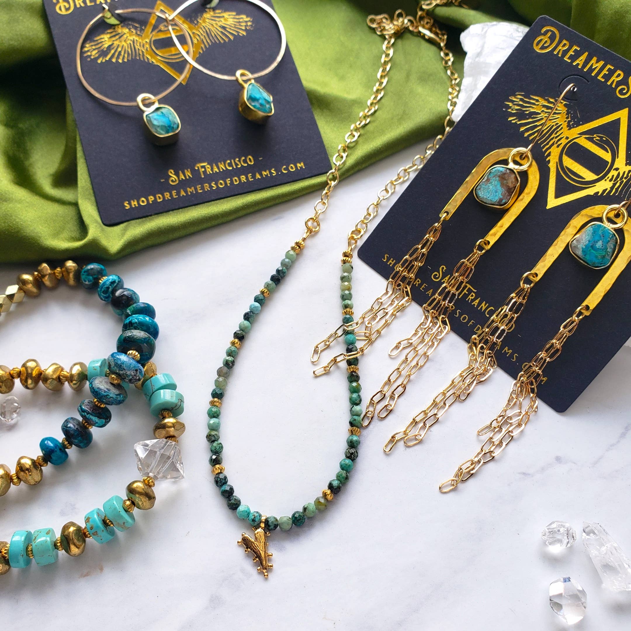 Handcrafted bohemian jewelry on a white surface: Dreamers of Dreams Chrysocolla Beaded Heart Charm Necklace, turquoise bracelets, and gold earrings with turquoise stones.
