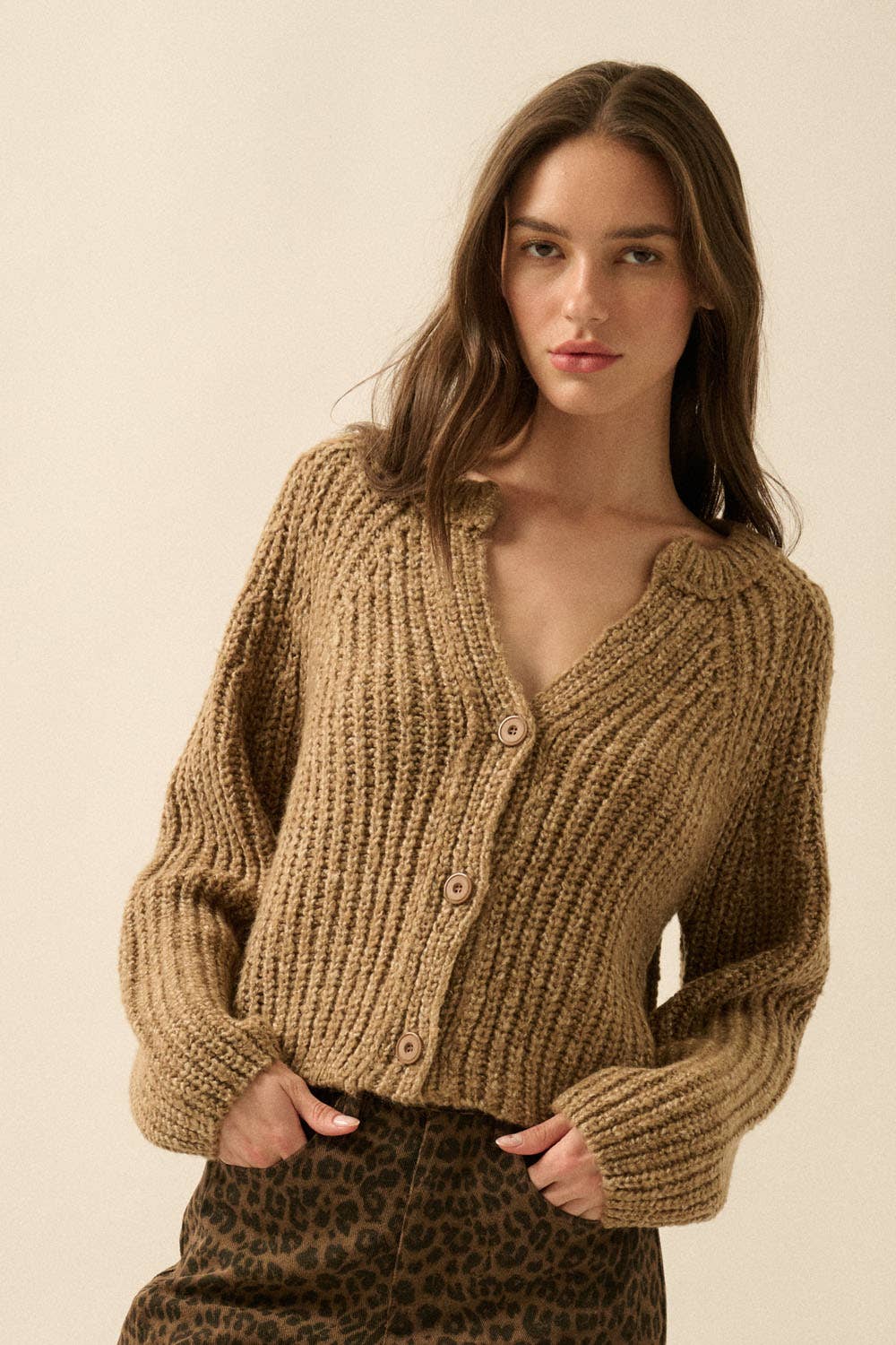 A woman with long brown hair wears the Promesa USA Confetti Button-Front Cardigan Sweater, hands in the pockets of leopard-print pants, standing against a plain light background.