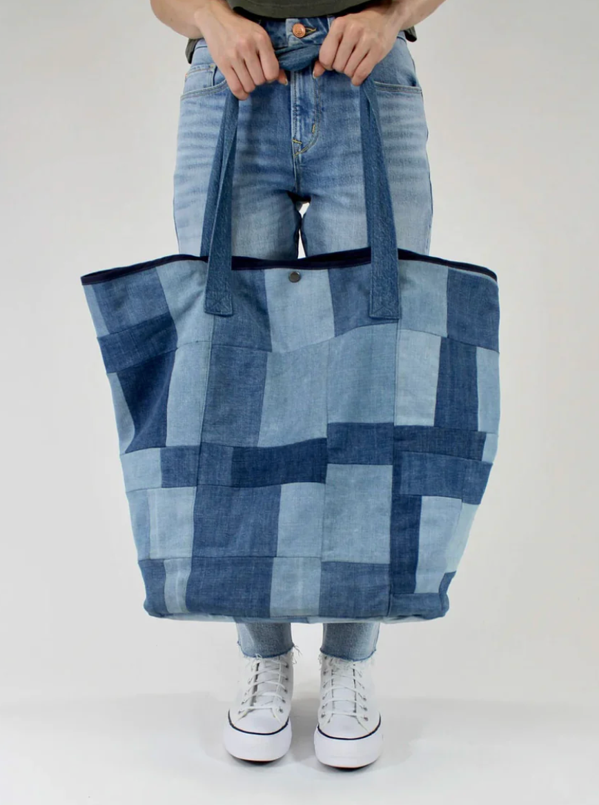 A person stands against a plain background, holding the Porter Blue Apparel PATCHWORK TOTE by its handles while wearing light blue jeans and white sneakers.