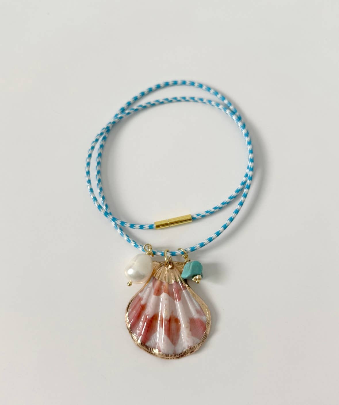 The Islas Necklace by Tramps+Thieves features a blue and white braided coastal design with a pink-spotted seashell pendant, white freshwater pearl, small turquoise stone, and gold accents, displayed on a white background.