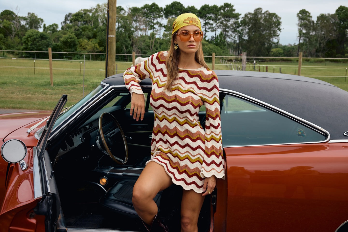 Woman in retro wavy striped dress and headscarf stands beside open door of vintage orange car outdoors