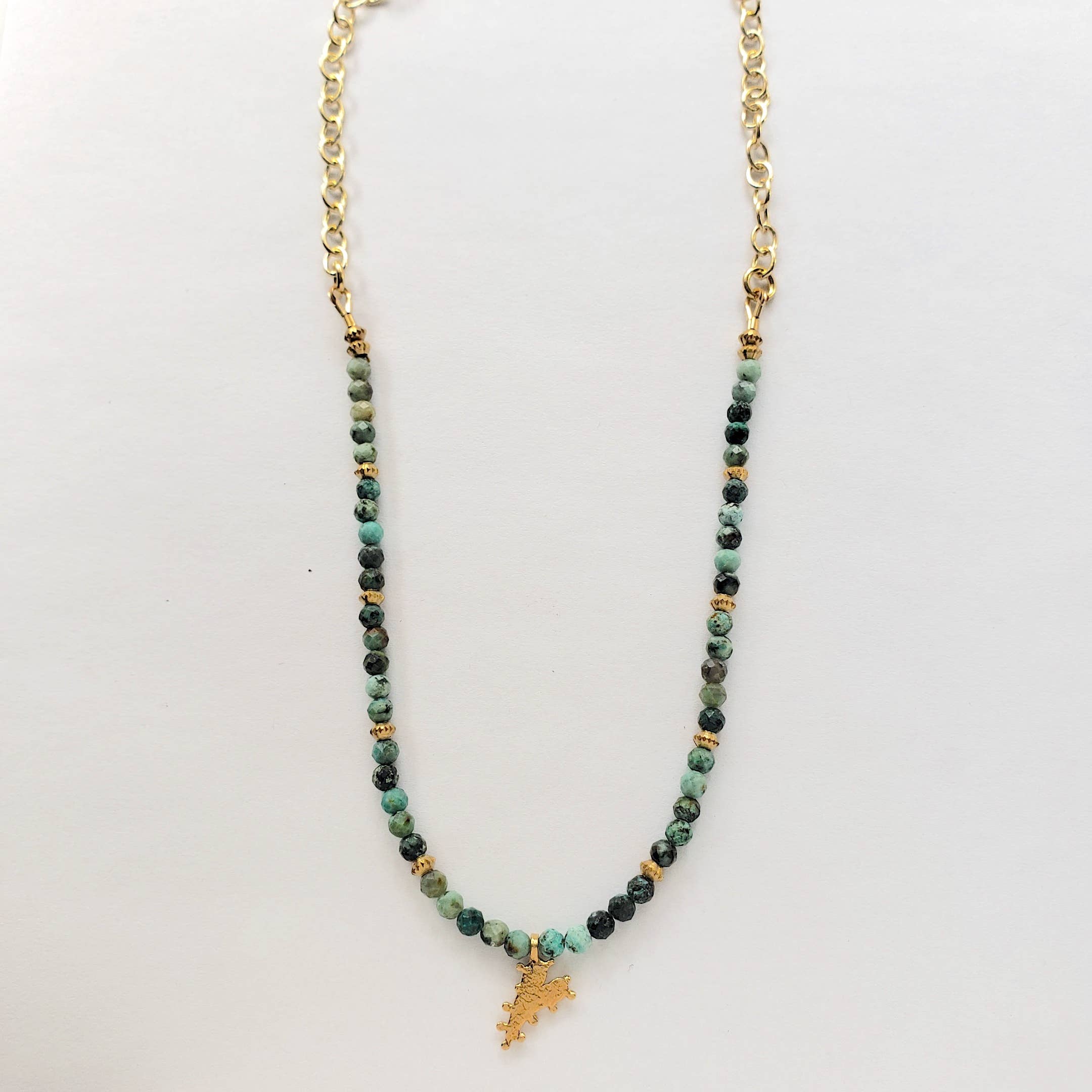 The Chrysocolla Beaded Heart Charm Necklace by Dreamers of Dreams features green beads, gold spacers, and a heart charm centerpiece. This bohemian piece is displayed against a plain white background.