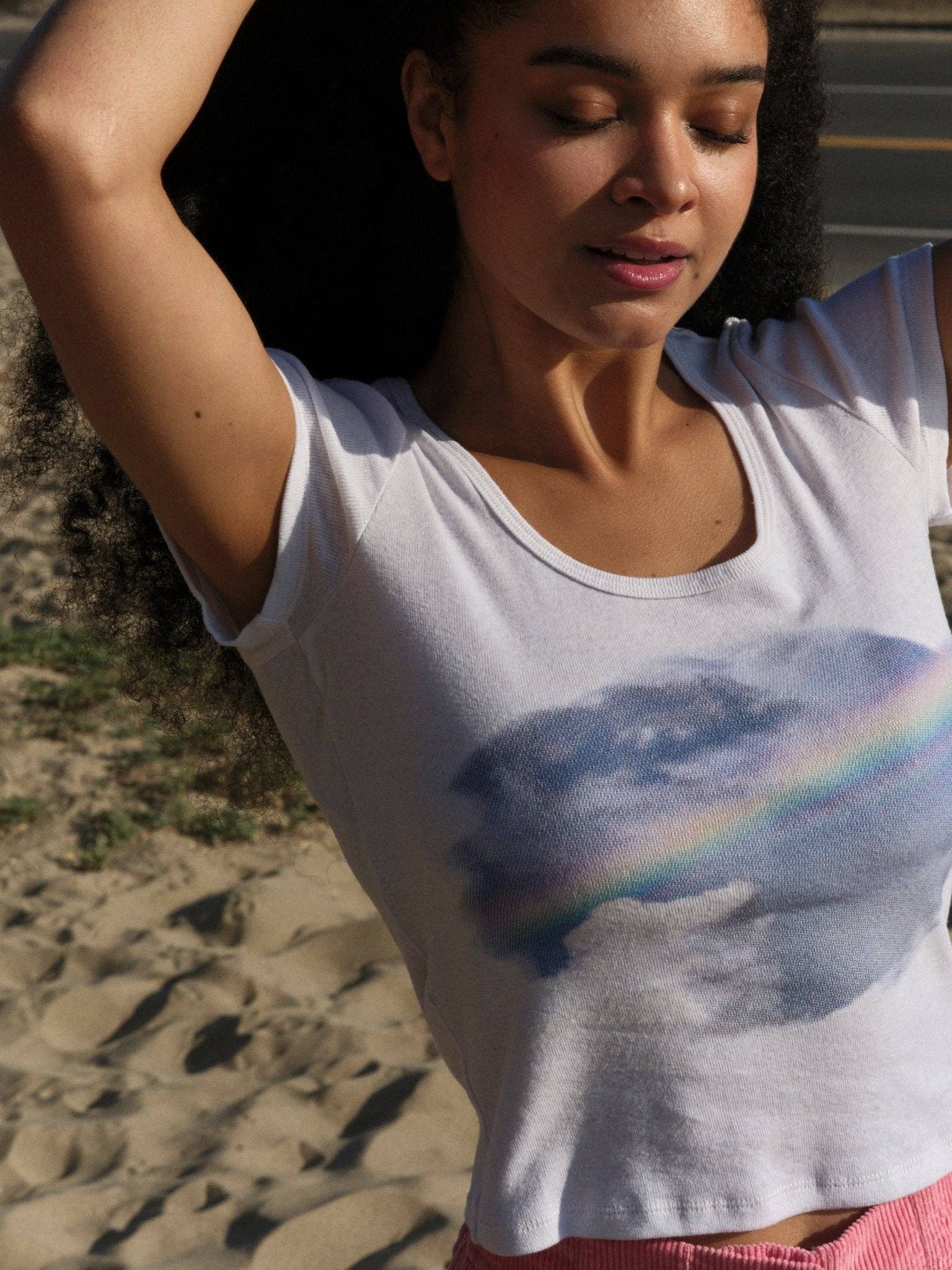 A woman with curly hair stands outdoors, eyes closed and arms raised in bliss. She wears the Mollusk CloudBreak Tee—a white shirt with a face and rainbow design inspired by California surf culture. Sandy ground and a road are in the background.