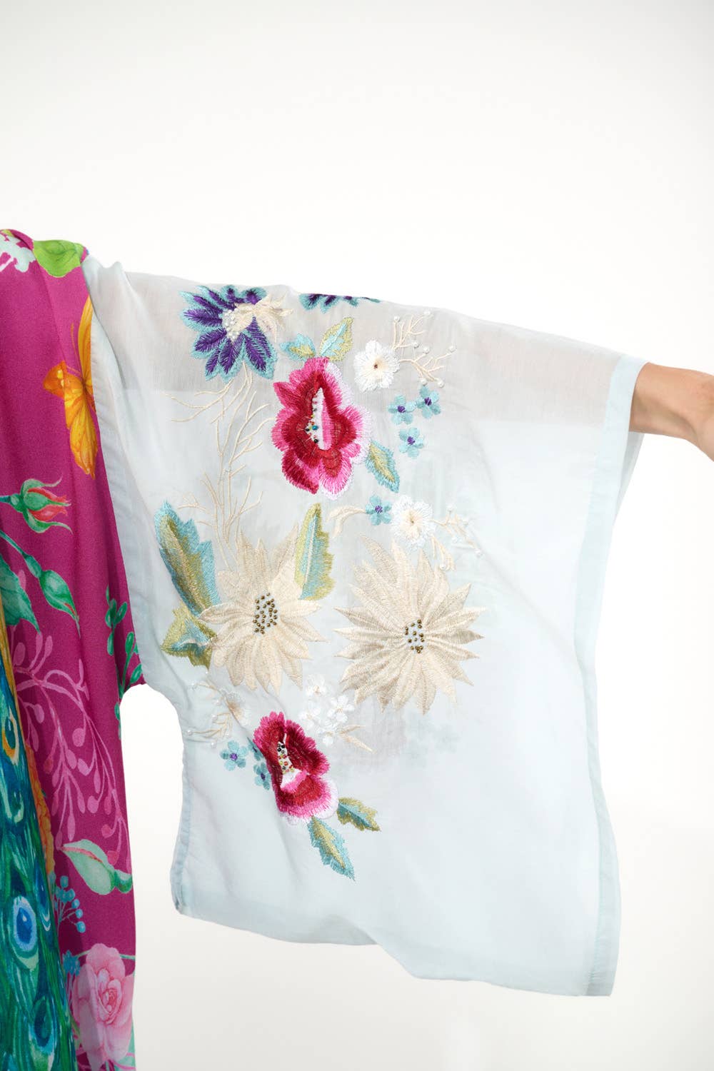 A person’s outstretched arm in a sheer light blue embroidered sleeve is beside the vibrant Marie Antoinette Kimono by Aratta, featuring colorful floral and peacock motifs.