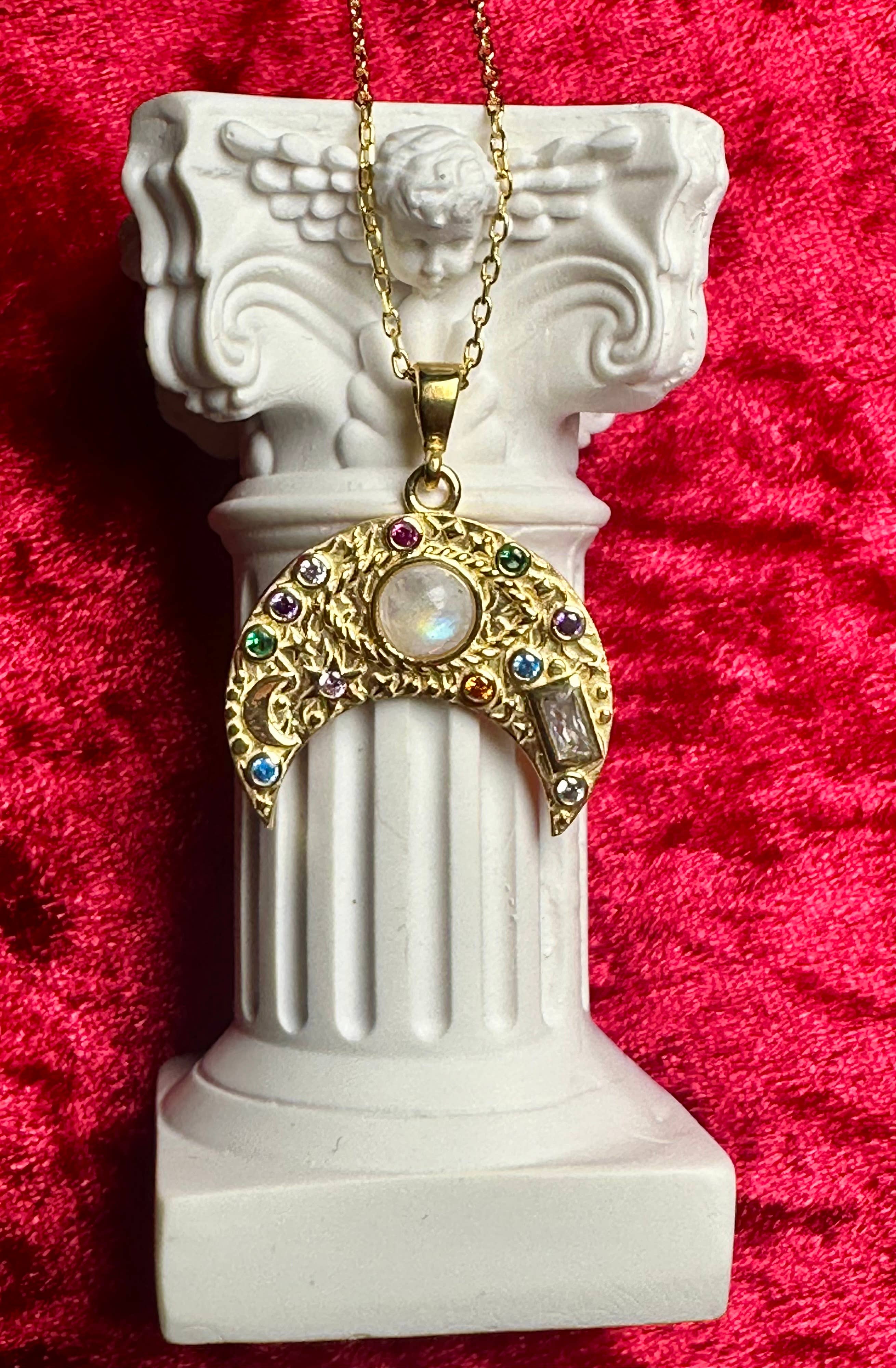 The Flesh & Soul CRESCENT MOON MOONSTONE Evil Eye Sterling Silver Necklace, featuring a crescent moon with vibrant gems and a central opal, is elegantly draped on a white angel-motif column against deep red velvet.