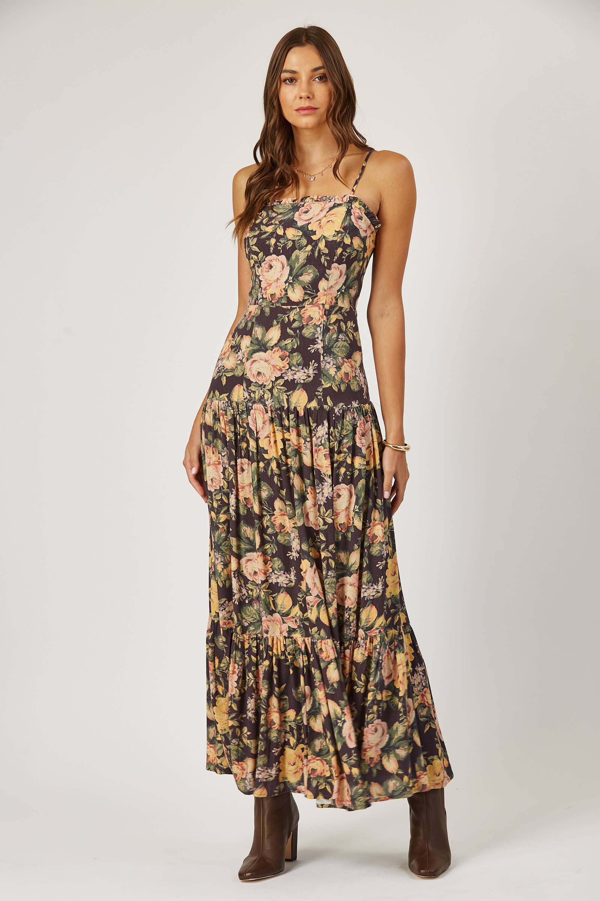 A woman stands against a plain background wearing the Isla Maude Priscilla laced back floral maxi dress with a flared skirt. Her hair is loose, and she accessorizes with brown ankle boots and a bracelet.