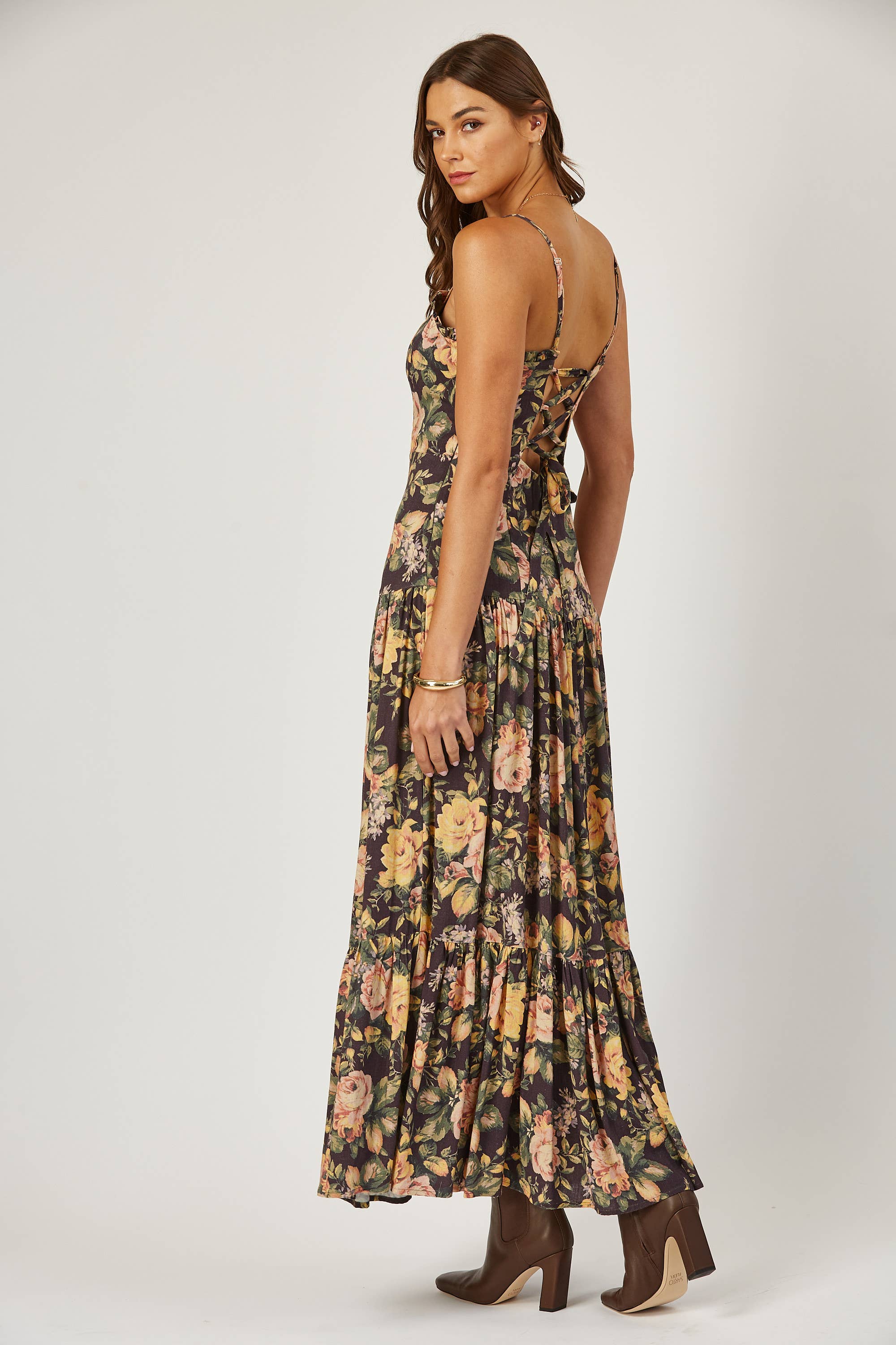 A woman in the Isla Maude Priscilla laced back floral maxi dress and brown ankle boots stands sideways, glancing over her shoulder against a plain light background.