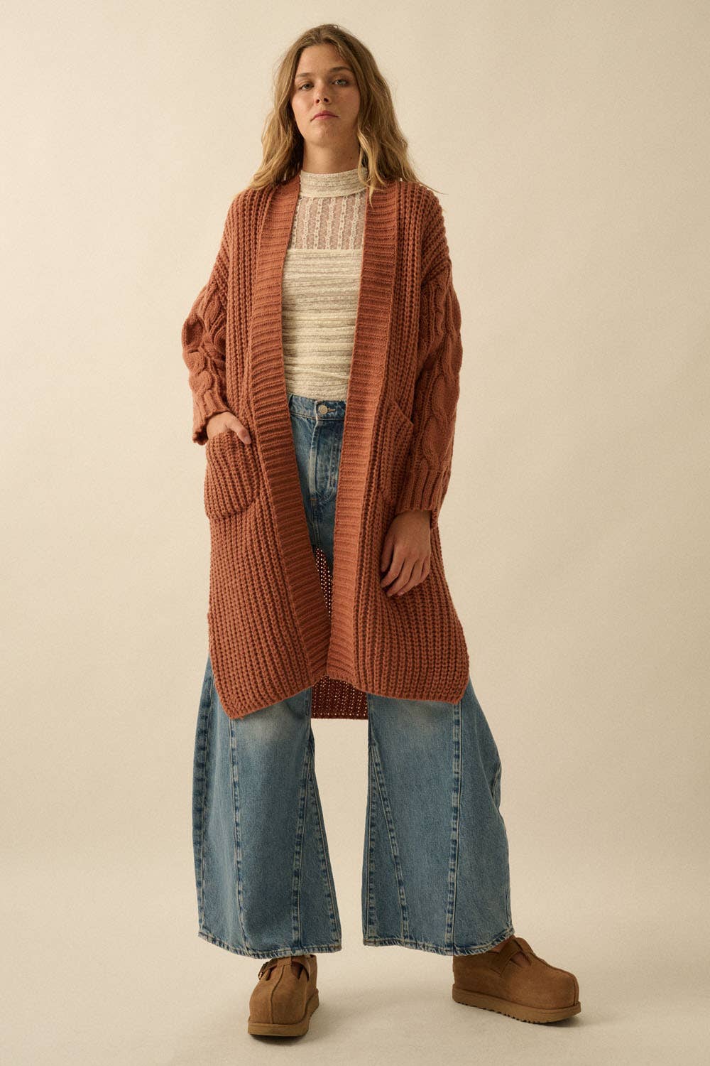 A woman wears the Promesa USA Solid Rib Knit Open Front Cable Knit Cardigan in rust over a cream knit top, paired with wide-leg blue jeans and tan platform shoes, posing against a plain beige background.