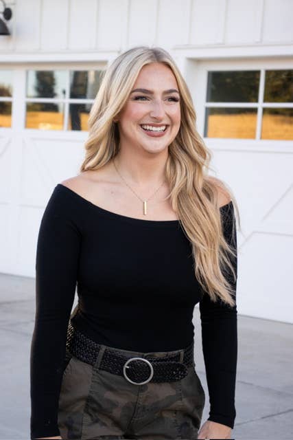A woman with long blonde hair stands outdoors, smiling in a black off-the-shoulder top and camo pants, accessorized with the Ginger Blue Classic Wide Braided Leather Belt in Black. White garage doors and sunlight are behind her.