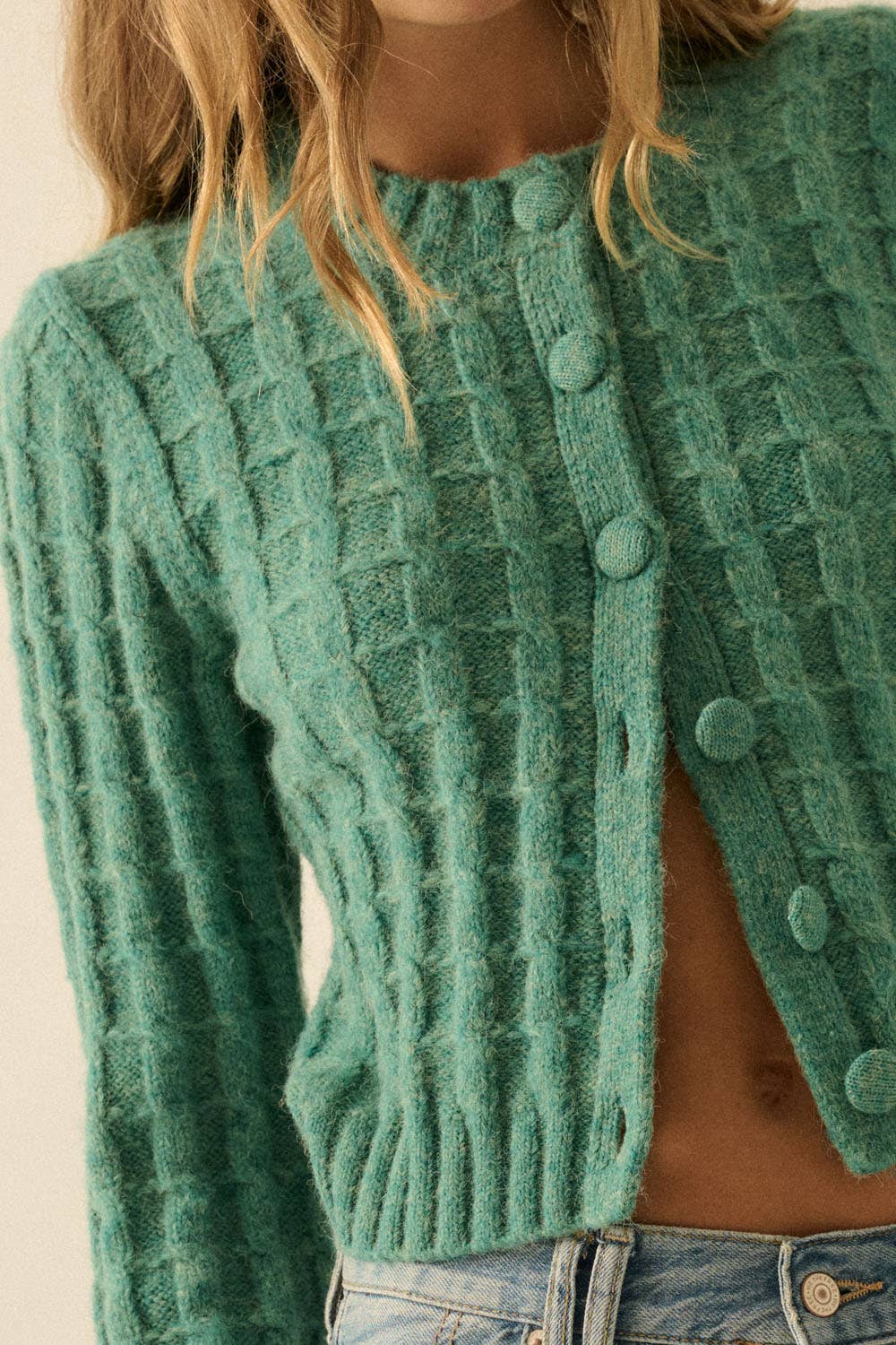 A person wears a Promesa USA Cable Knit Button-Up Cardigan Sweater in textured teal-green, partially unbuttoned to show the midriff, paired with light blue jeans. The person's face is not shown.