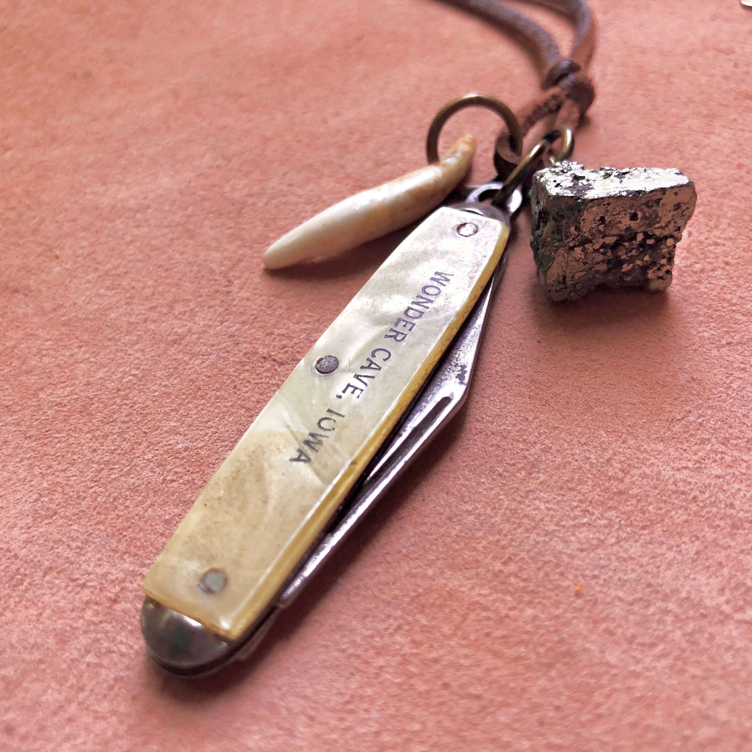 The ASTALI Vintage Pocket Knife & Coyote Tooth Necklace features a WONDER CAVE, IOWA-engraved knife, pyrite nugget, and deerskin leather cord, beautifully displayed on pink fabric.