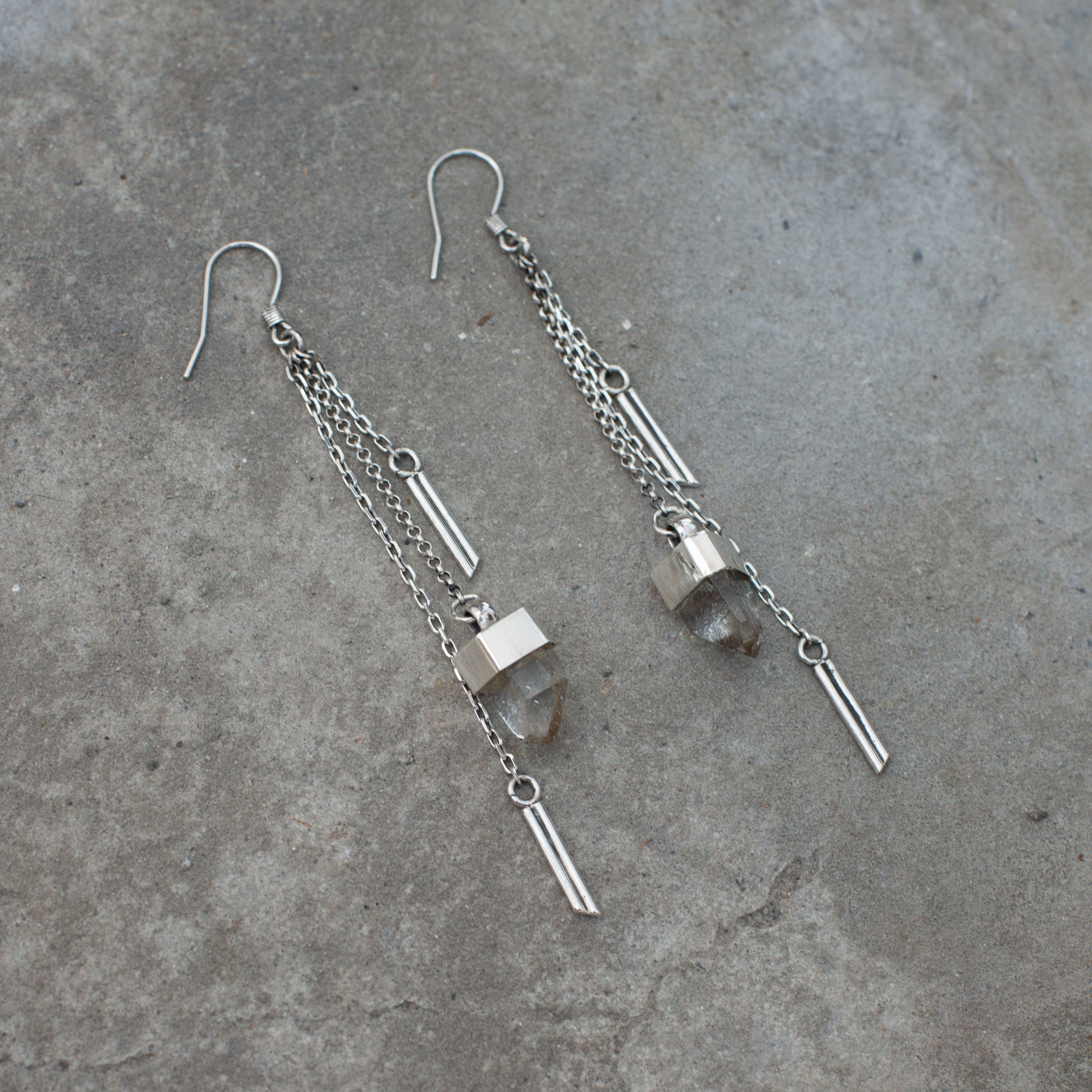 The Jan Hilmer + Sparrow Crystal dangle earrings—long sterling silver with multiple chains, geometric accents, and clear crystal pendants—are shown on a textured gray surface.