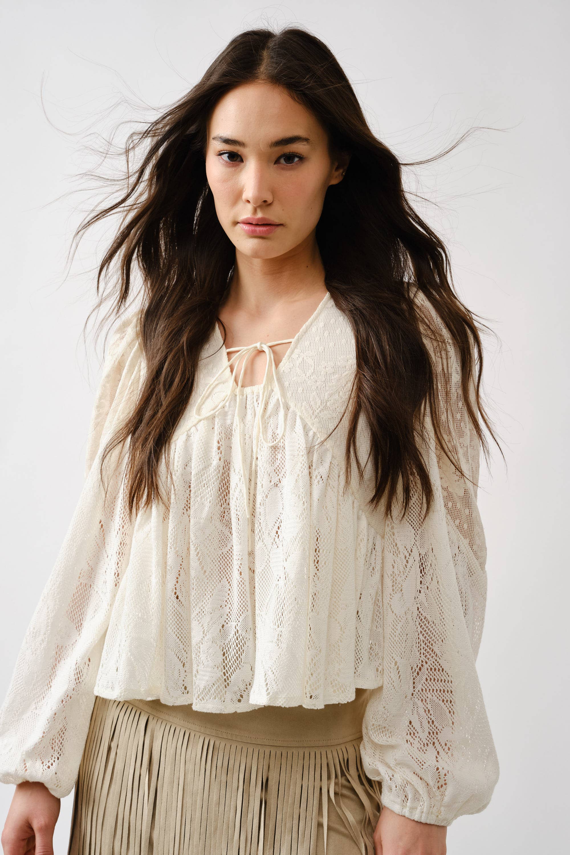 A woman with long, wavy dark hair wears the Aaron & Amber Long Sleeve Lace Babydoll—a loose, cream floral lace top with a neckline tie—paired with a light beige fringed skirt, standing against a plain white background.