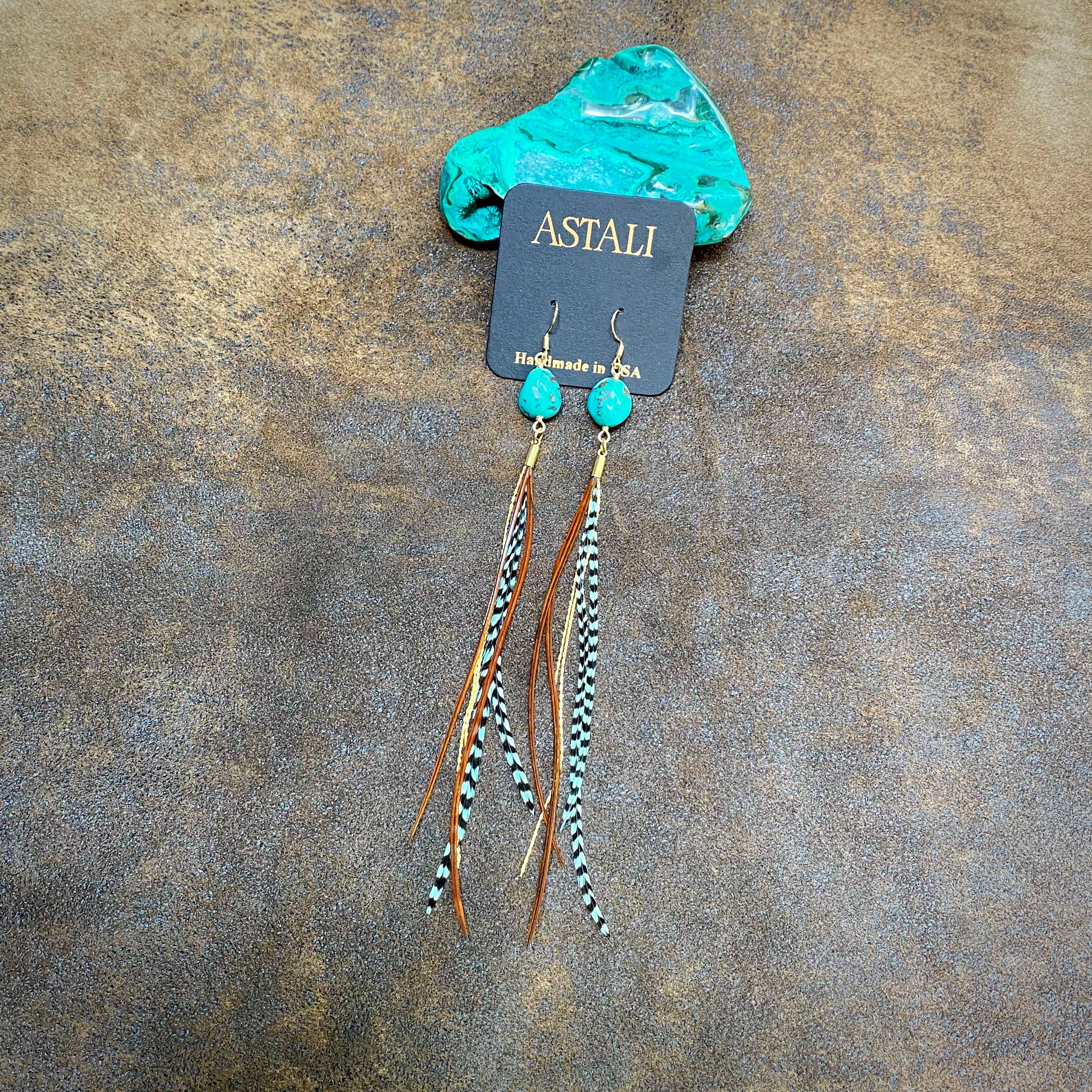 The Turquoise & Mini Feather Earrings - Teal Grizzly & Fawn/Gold by ASTALI are shown on a branded jewelry card, featuring a turquoise stone and dangling brown, black, and white feathers against a textured brown background.