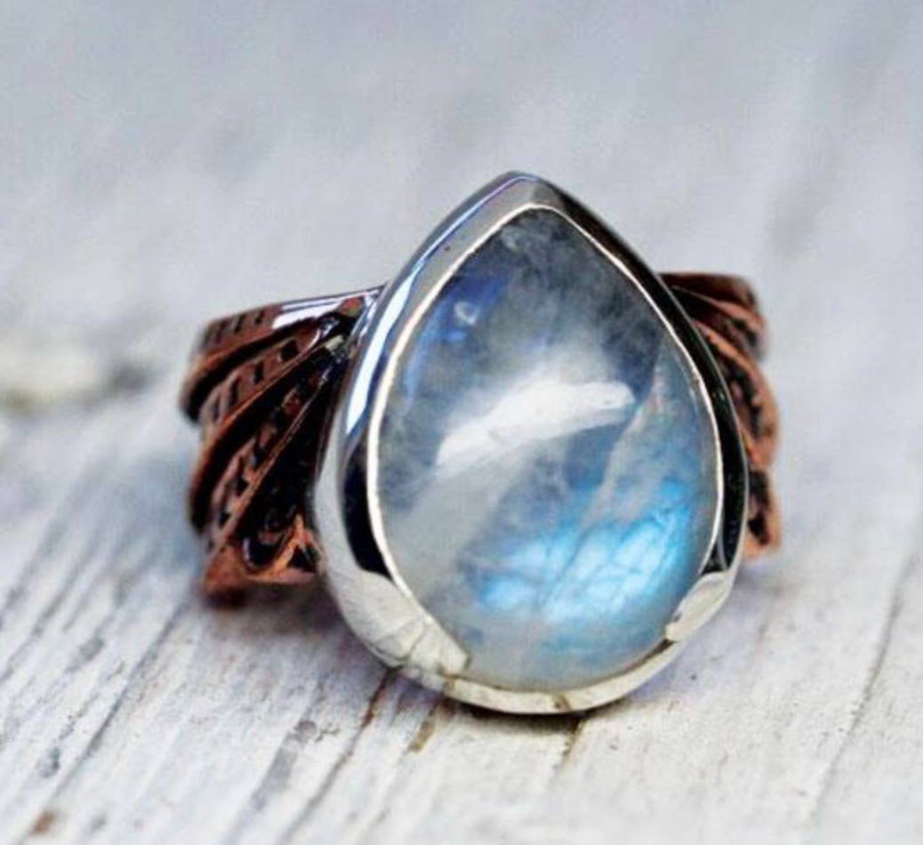 The Winged Ring by bahgsu jewels features a large, teardrop moonstone set in silver atop an intricate copper band, elegantly displayed on a light wooden surface.