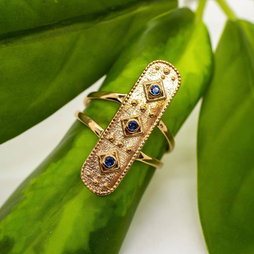 The Baizaar Sapphire Healer Ring features an elongated gold brass design with textured details and three faceted sapphires, shown resting on a green leaf in gentle light.
