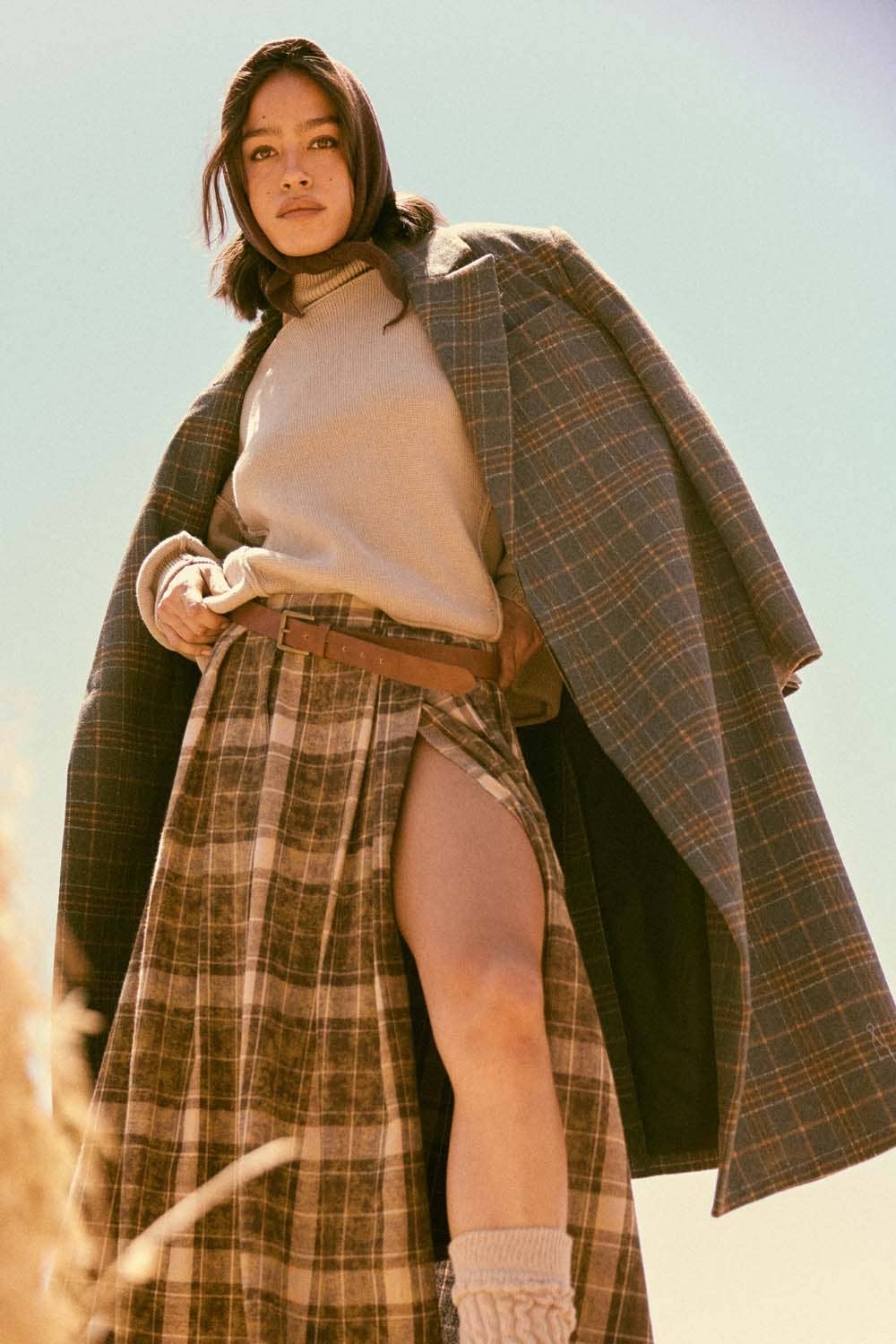 A woman stands outdoors in sunlight, wearing a beige turtleneck and plaid skirt with a high slit. She confidently looks at the camera with the Plaid Felt Double-Breasted Calf-Length Overcoat by Promesa USA draped over her shoulders.