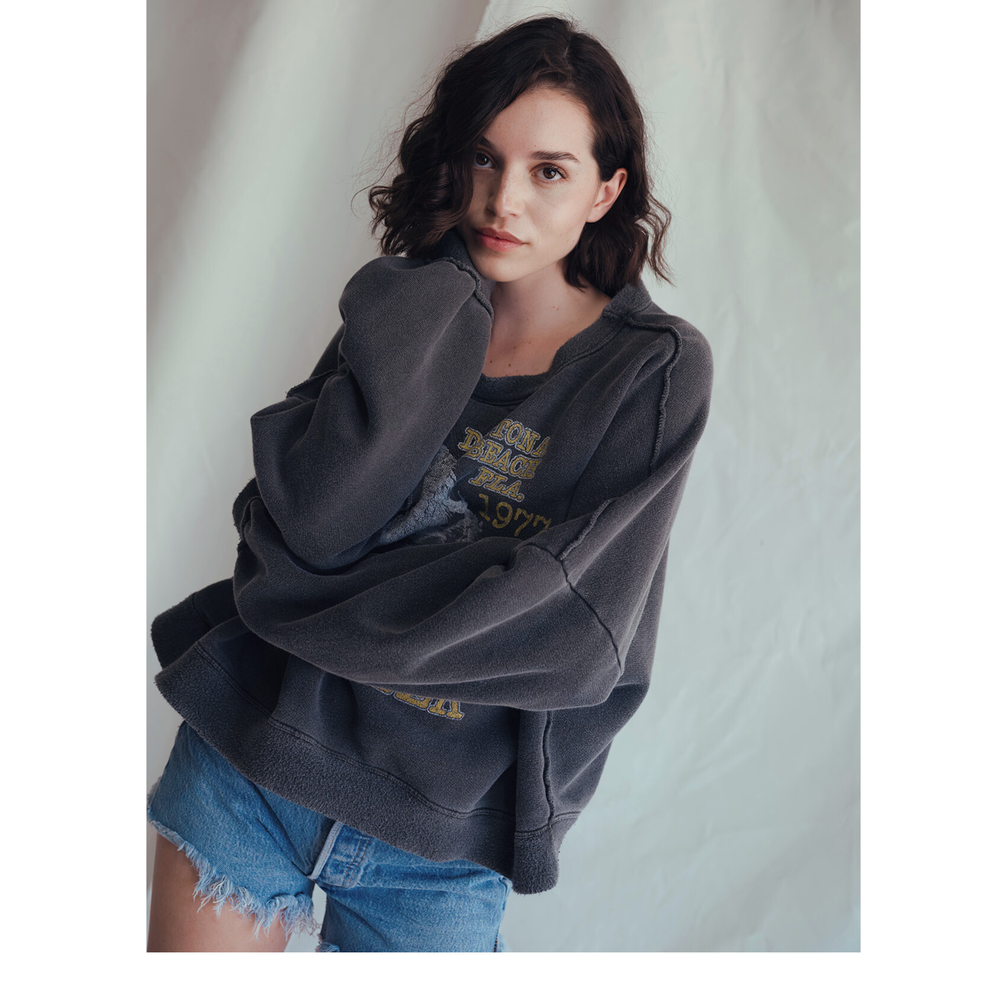 A young woman with wavy dark hair wears the People Of Leisure Daytona Sweatshirt, featuring a vintage eagle graphic, and denim shorts as she stands before a light background, looking at the camera with a neutral expression.