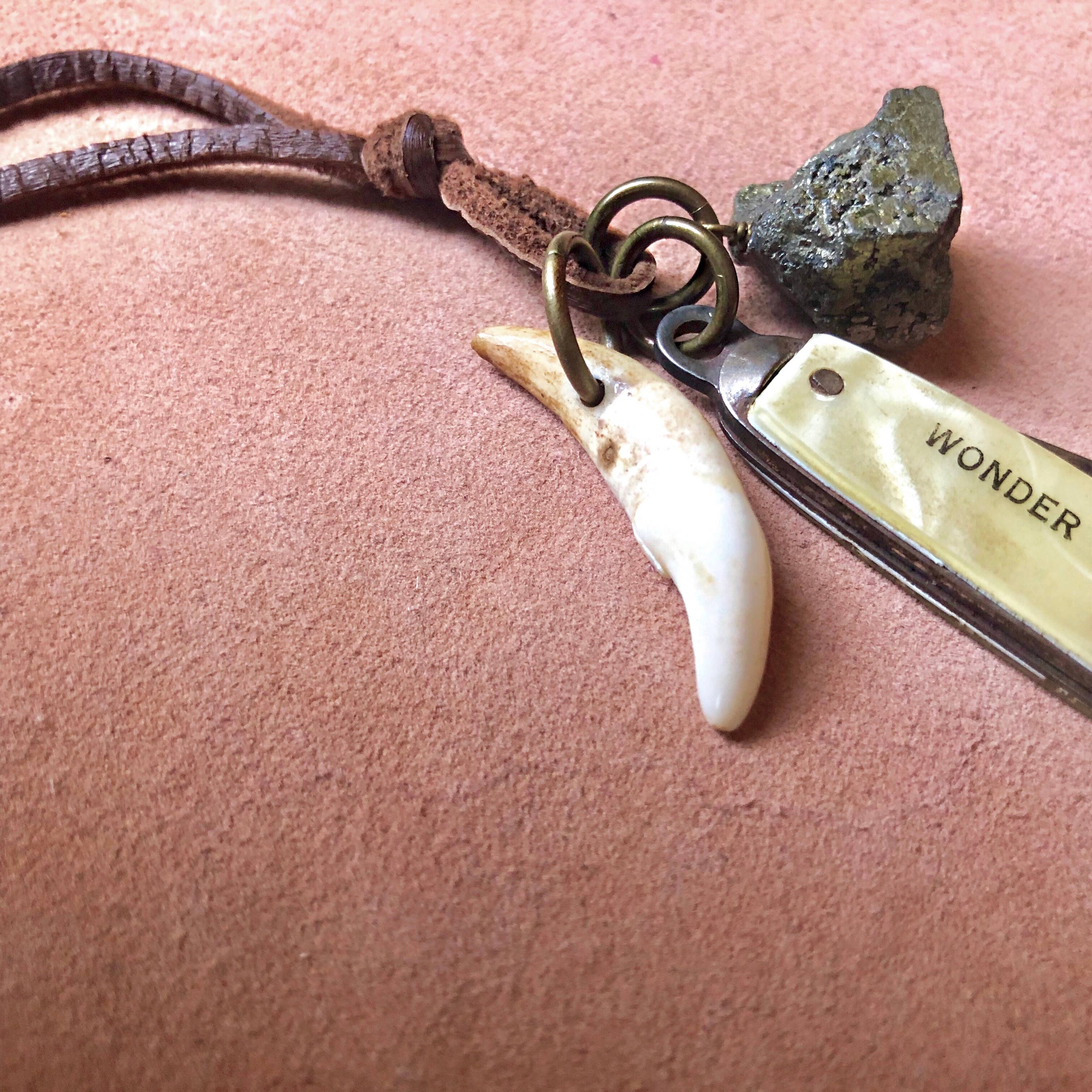 The ASTALI Vintage Pocket Knife & Coyote Tooth Necklace features a deerskin leather cord, pyrite stone, engraved vintage knife pendant, and coyote tooth, all showcased on a soft pink surface.