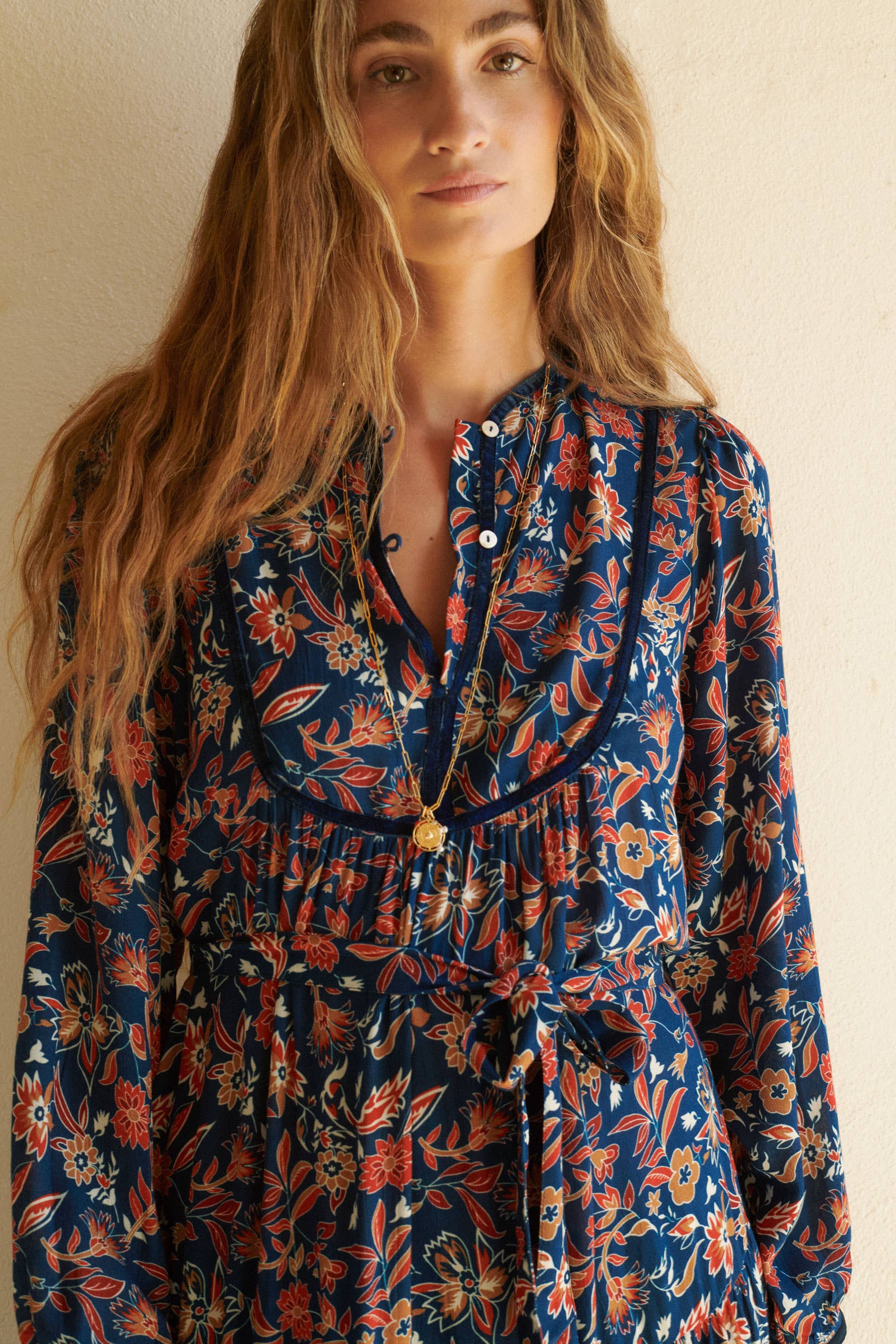 A woman with long wavy hair wears the Aura Dress Navy by Sonmer Shop USA—a floral autumn dress with a ruffled waist and buttoned, adjustable V-neckline. She accessorizes with two gold necklaces, standing against a light-colored wall.