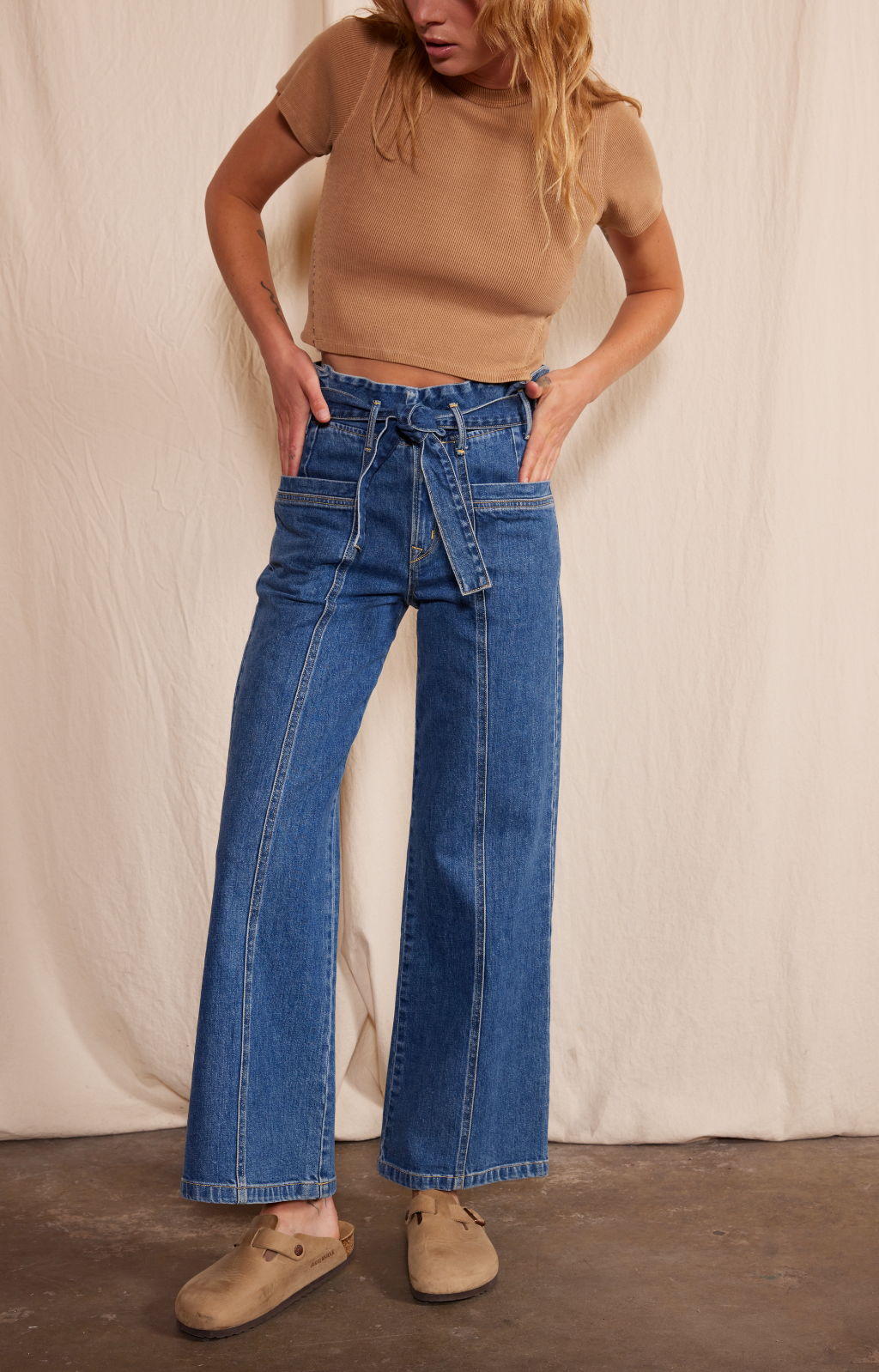 A woman stands against a beige fabric backdrop, wearing Porter Blue Apparel’s PENNY PAPERBAG WIDE LEG IN LAGUNA WASH—high-waisted organic cotton jeans—paired with a short-sleeve tan top and slip-on shoes. Her hands are in her pockets as she looks down.