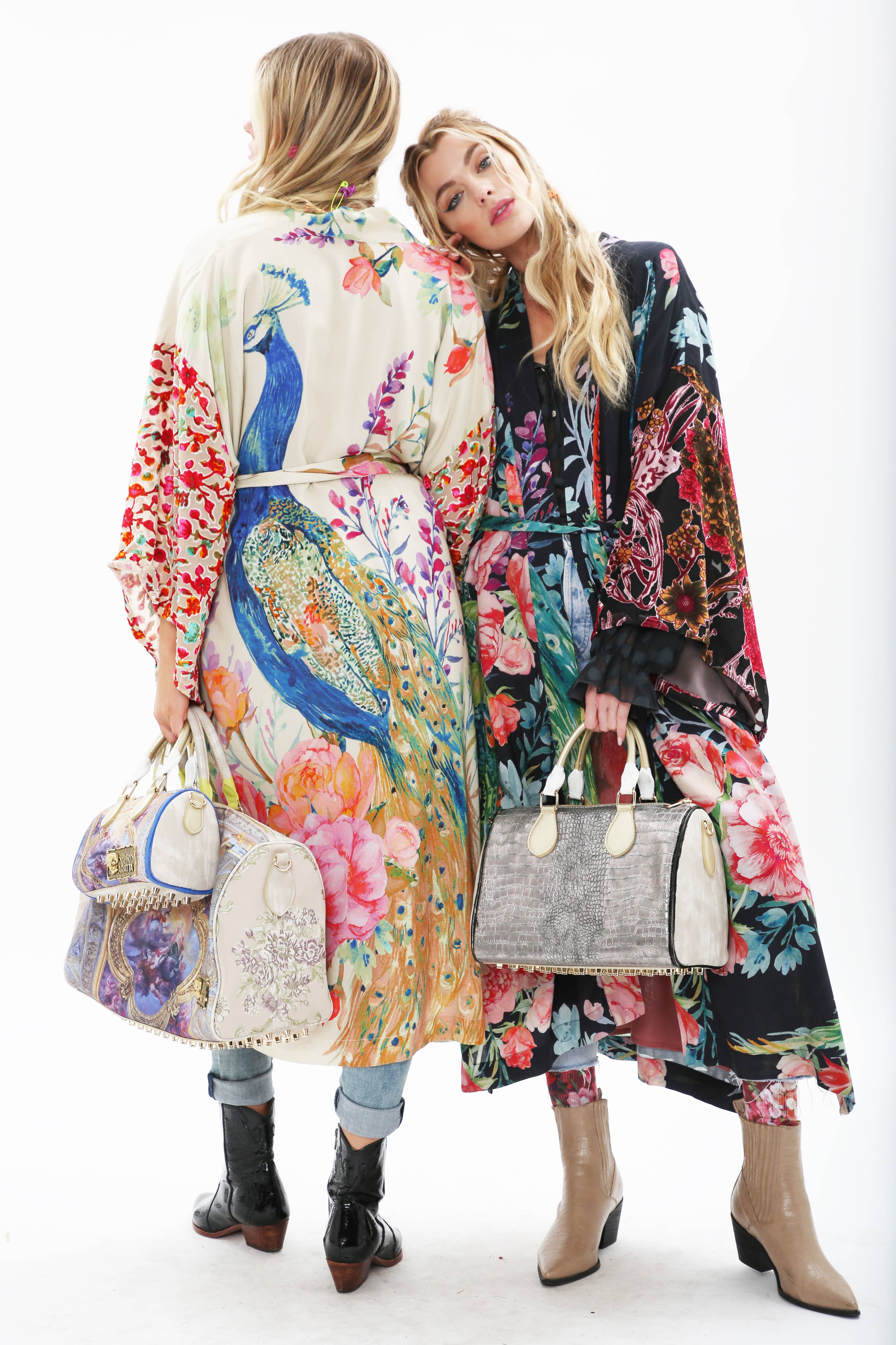 Two women stand back-to-back: one in Aratta's Sweet Fantasy Kimono with burnout velvet sleeves, the other in a lined, belted kimono featuring floral and geometric designs. Both carry large handbags and wear ankle boots, their loose waves completing the look.