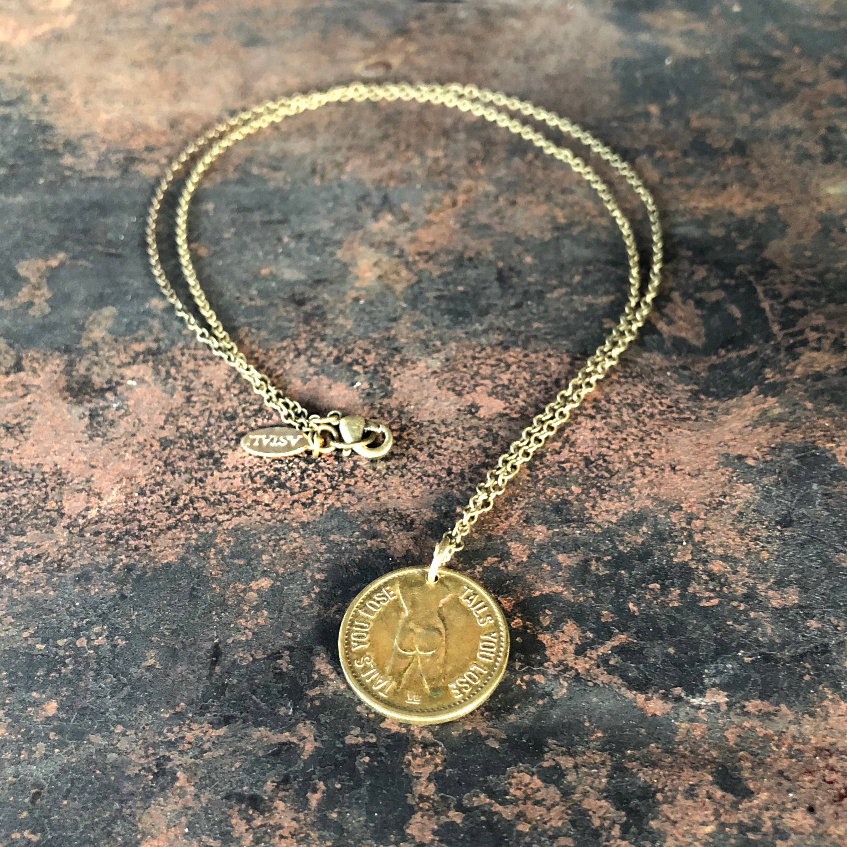 The ASTALI Vintage Heads & Tails Peepshow Token Coin Necklace is displayed on a textured brown and black surface—a standout collectible piece inspired by vintage peepshow tokens, featuring a round pendant engraved with ITALY.