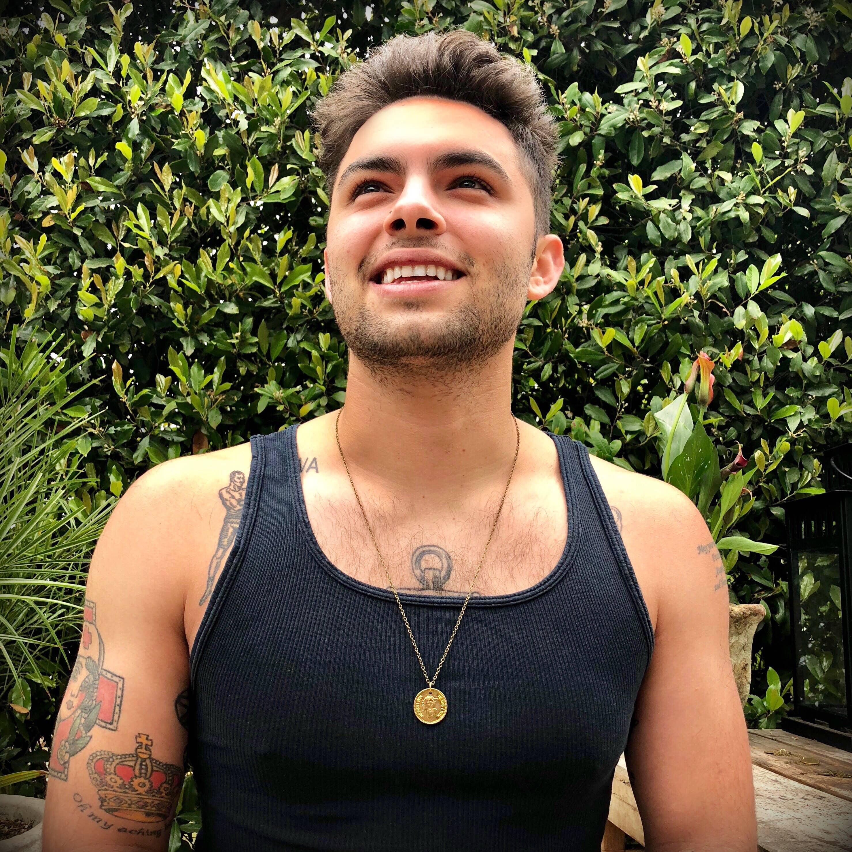 A smiling young man with short hair wears an ASTALI Vintage Heads & Tails Peepshow Token Coin Necklace and a black tank top, sitting outdoors in front of lush green plants. Tattoos decorate his arms and chest.
