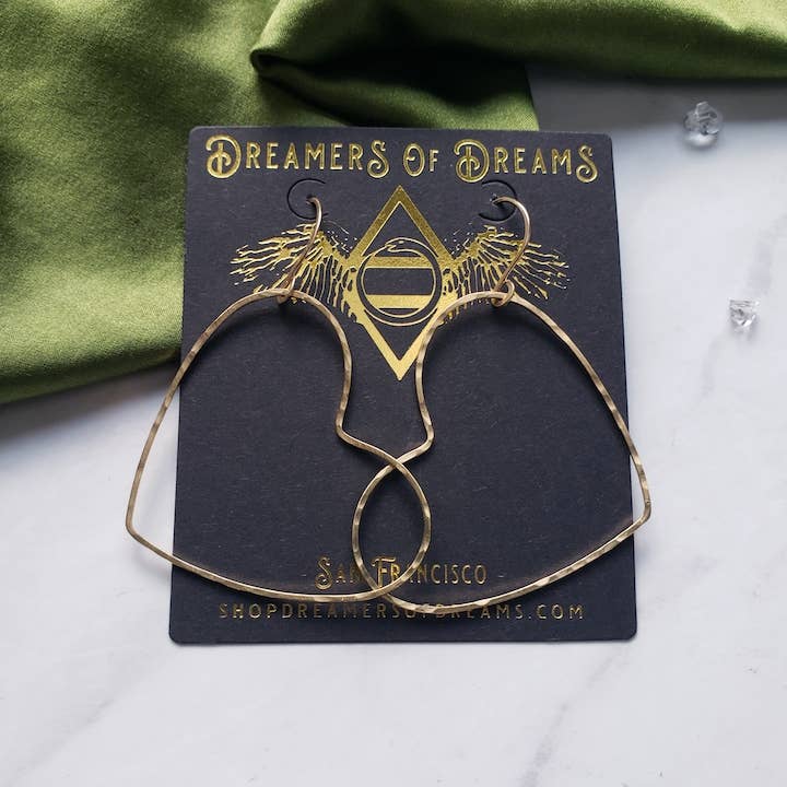 Big Gold Heart Hoop Earrings by Dreamers of Dreams displayed on a branded dark card, set on a white surface with green fabric and a clear gem in the background.