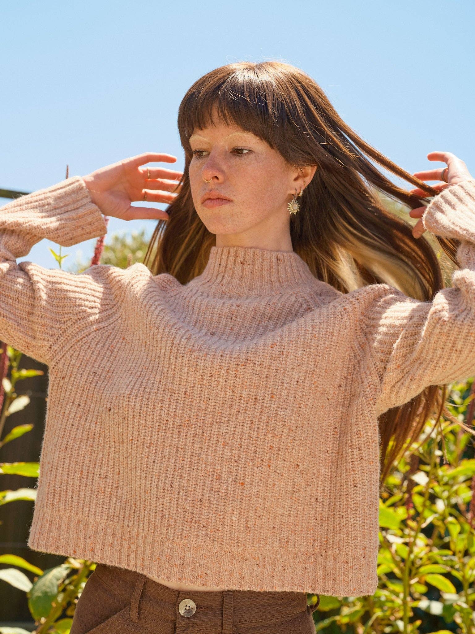 A woman with long brown hair stands outdoors under a clear blue sky, wearing the Mollusk Teddy Sweater in light pink with brown pants; she holds her hair with both hands, surrounded by lush greenery.