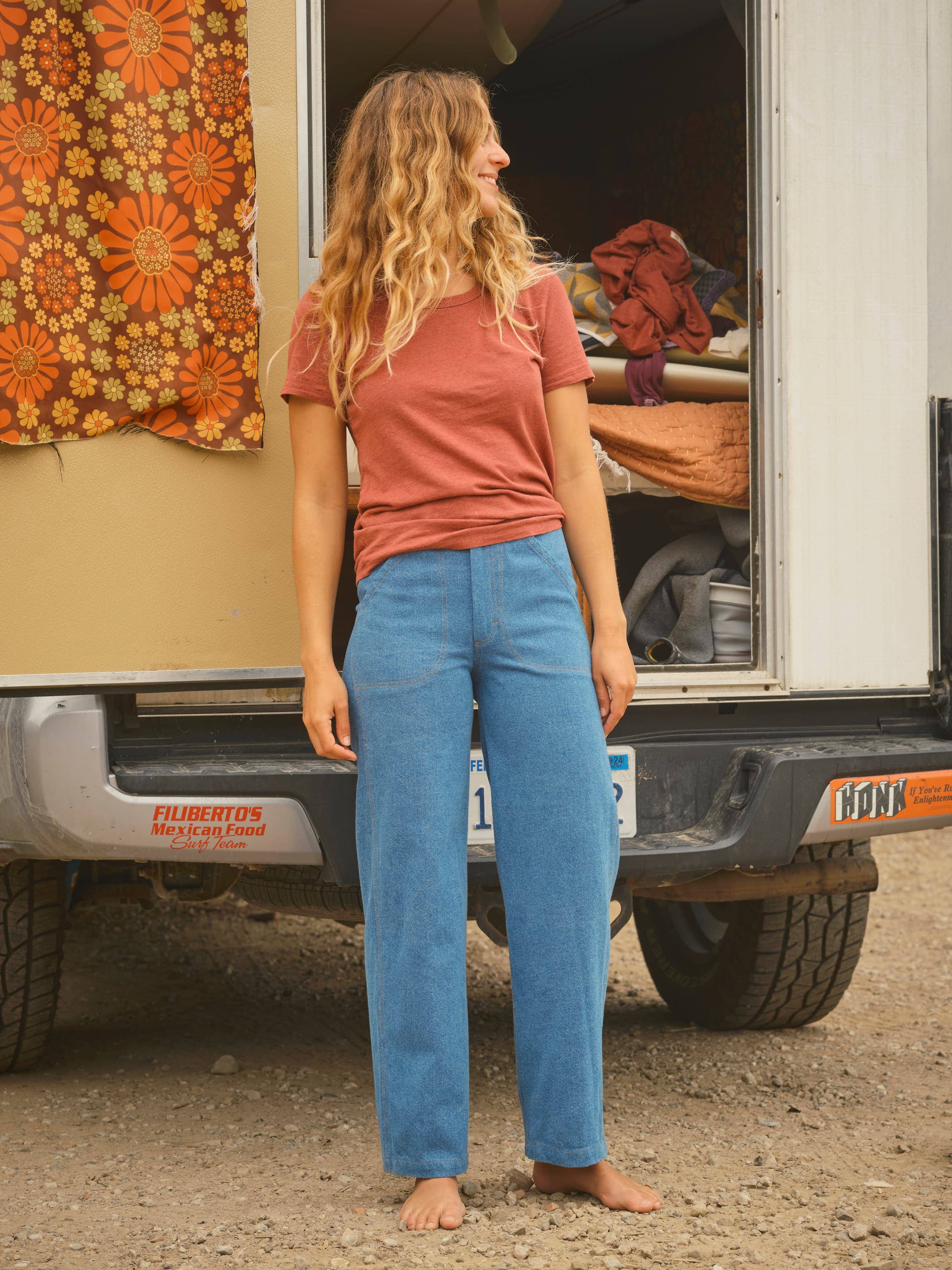 A woman with wavy blonde hair, in a rust t-shirt and Mollusk Painter Pants in Indigo from the women’s bottoms collection, stands barefoot outside an open vehicle of colorful clothes, looking left and smiling.