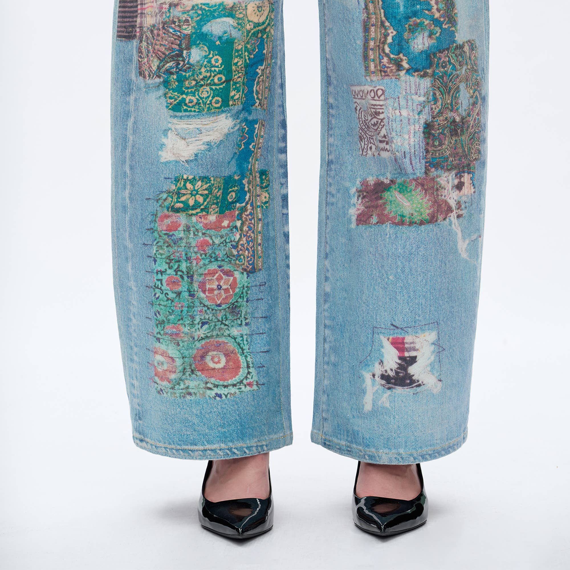 A close-up of lower legs in BAYEAS Light Blue Patch Pattern High-Rise Barrel Jeans, featuring comfort stretch denim and colorful patches, paired with black pointed-toe heels against a plain white background.