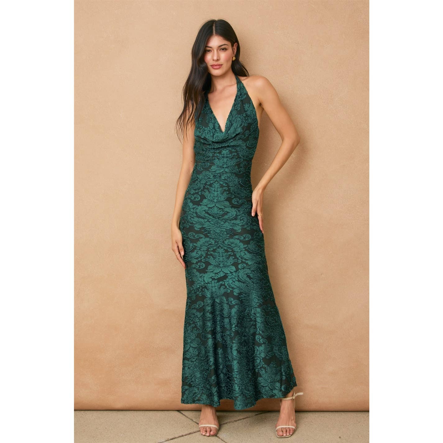 A woman with long, dark hair stands against a tan background wearing the Fame Draped Halter Neck Maxi Dress by Dress Forum in emerald velvet burnout with a subtle floral pattern, styled with strappy nude heels.