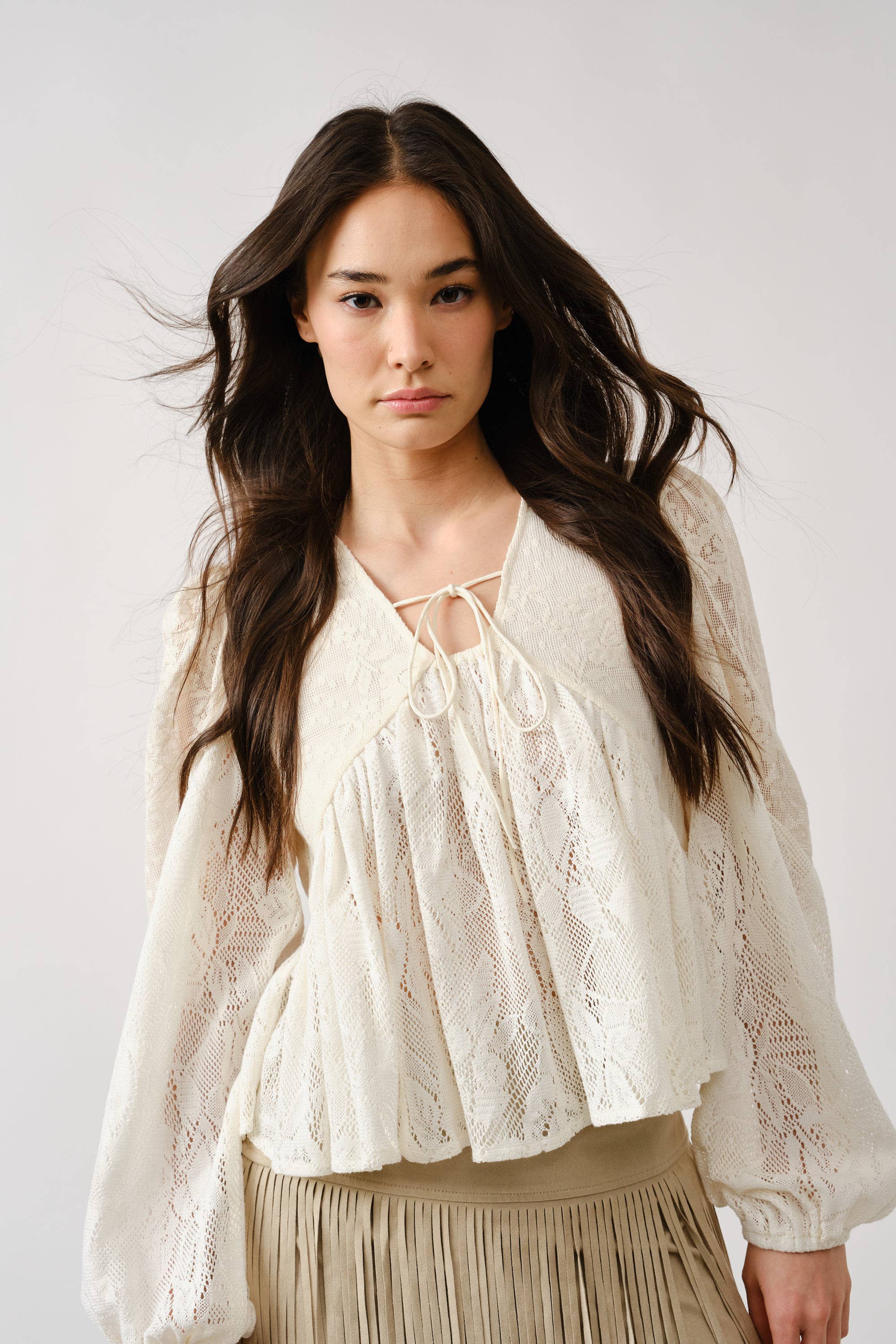A woman with long dark hair wears the Aaron & Amber Long Sleeve Lace Babydoll, a loose white floral lace blouse with a tie neckline, paired with a pleated beige skirt, standing against a neutral background.