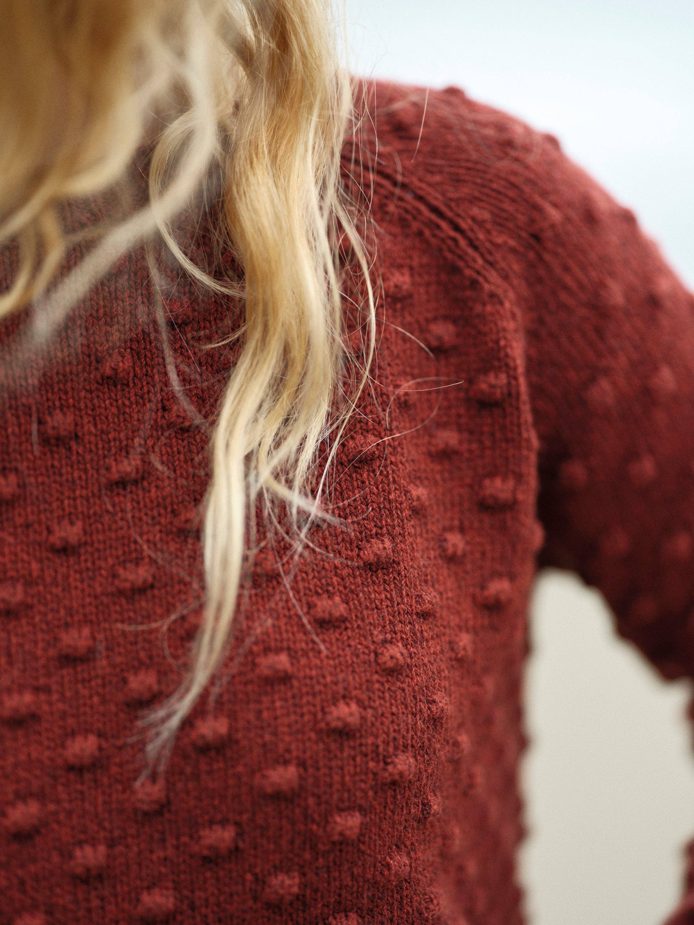 A close-up of a person wearing the Mollusk Pearl Sweater, a vintage-inspired dark red top with raised polka dots. Wavy blonde hair drapes over their shoulder.