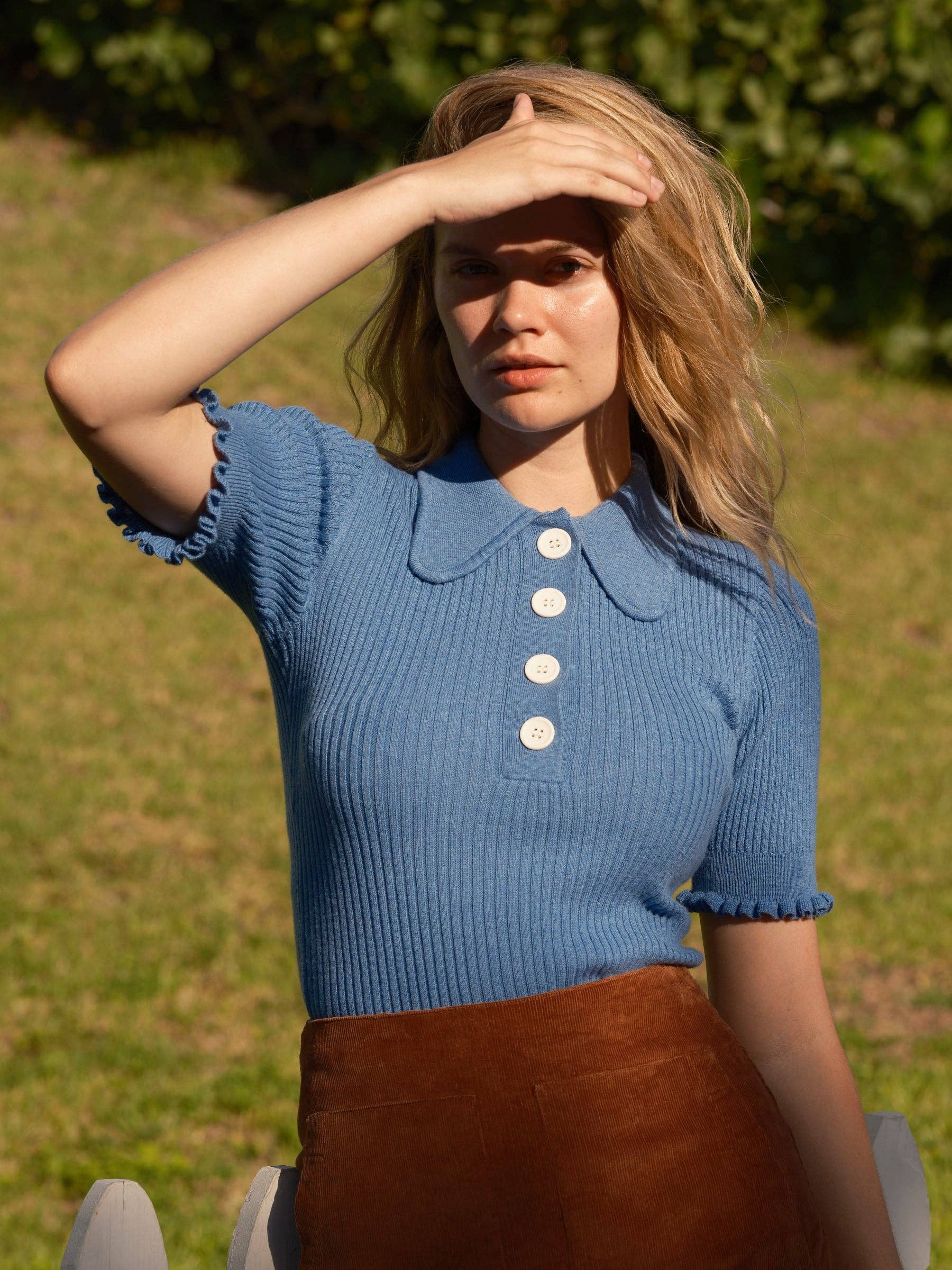 A woman outdoors in sunlight wears the Mollusk Belle Tee, a vintage-inspired blue ribbed top with large white buttons, paired with a brown skirt. She shields her eyes from the sun, greenery in the background.