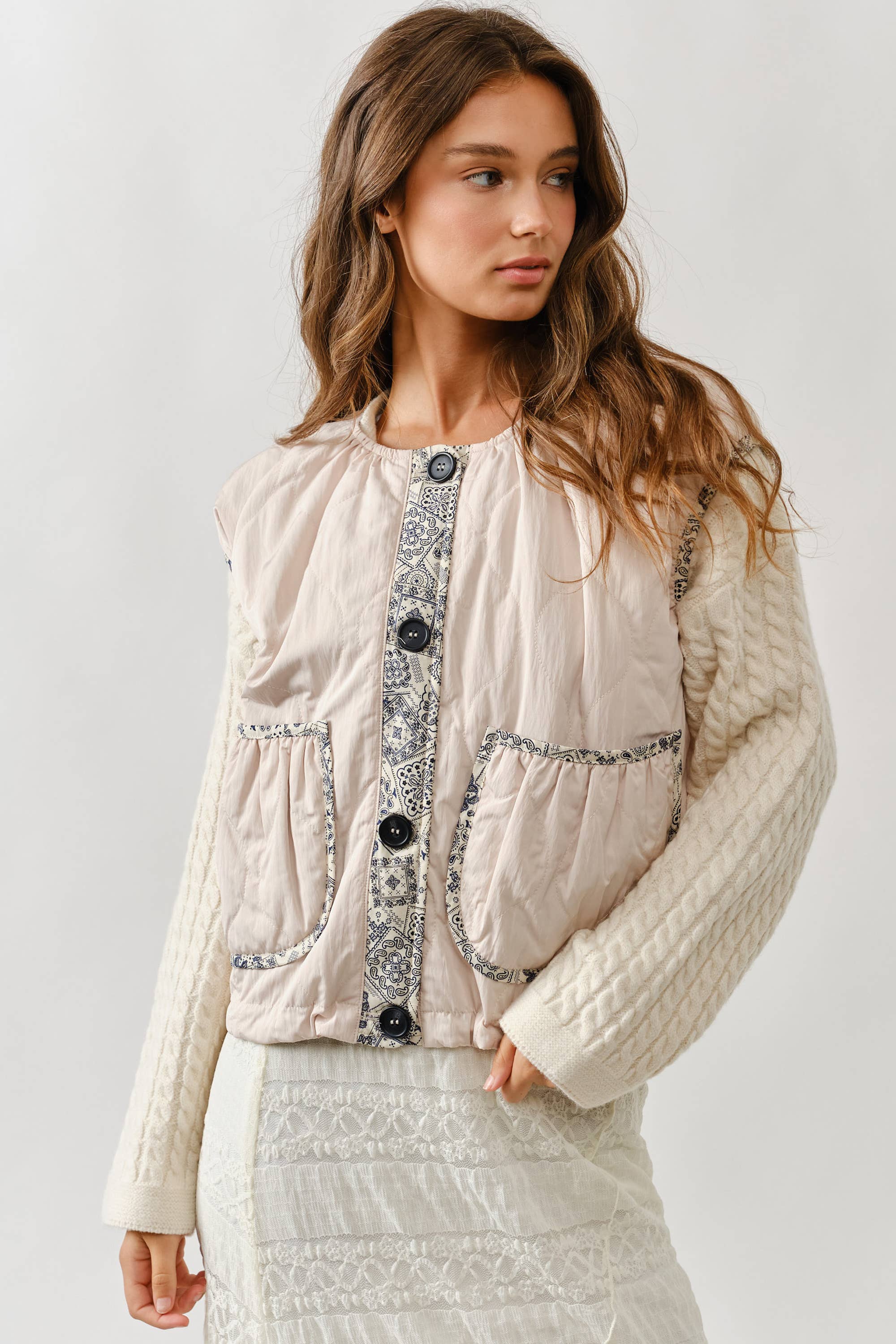 A woman wears the Aaron & Amber Contrast Detail Lightweight Vest, featuring patterned trim and large buttons, layered over a cream cable-knit sweater and white lace skirt, standing against a plain light background.