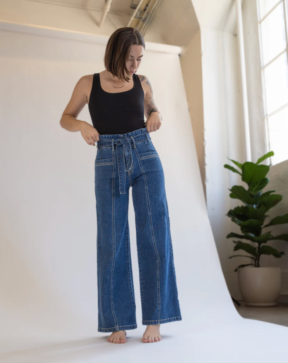 A woman stands barefoot on a studio set, wearing Porter Blue Apparels PENNY PAPERBAG WIDE LEG IN LAGUNA WASH jeans and a black tank top. A green potted plant by the window adds a natural touch to the background.