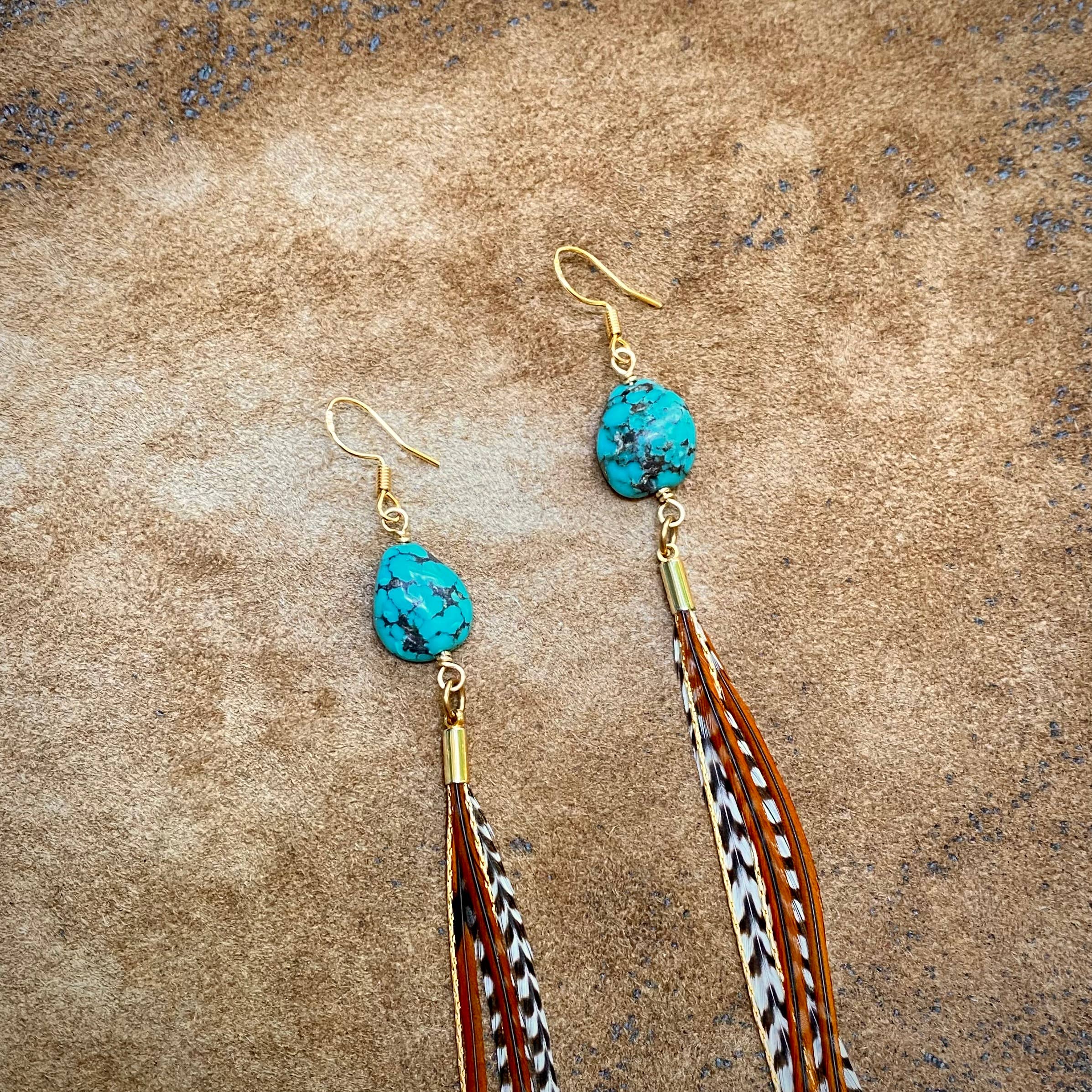 The ASTALI Turquoise & Mini Feather Earrings - Grizzly & Fawn/Gold feature gold-plated hooks and long feather strands in brown, black, and white, shown on a textured tan surface.