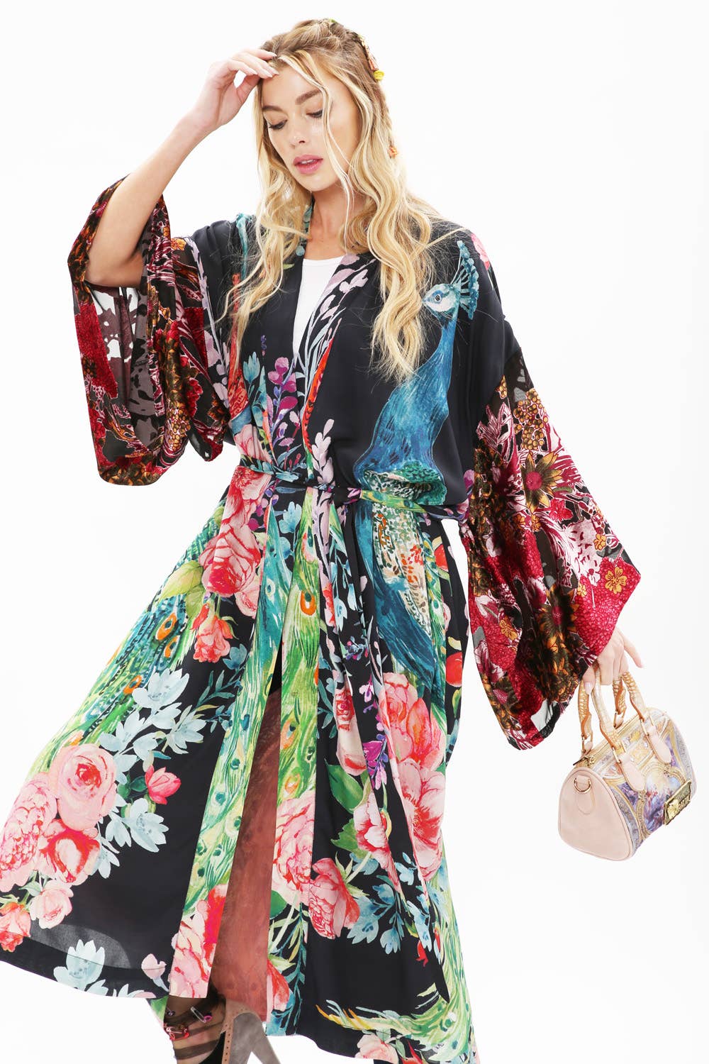 A woman in the Aratta Sweet Fantasy Kimono with wide sleeves and peacock floral print holds a small beige handbag. She has long, wavy blonde hair and stands against a white background.
