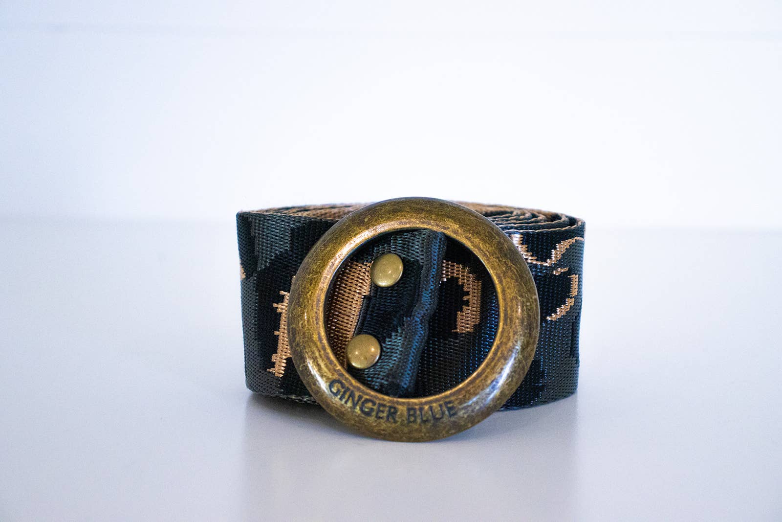 A rolled-up fabric belt with a large round cinch buckle engraved Ginger Blue sits on a white surface. The Wild Child Leopard features a dark blue and tan leopard print pattern.