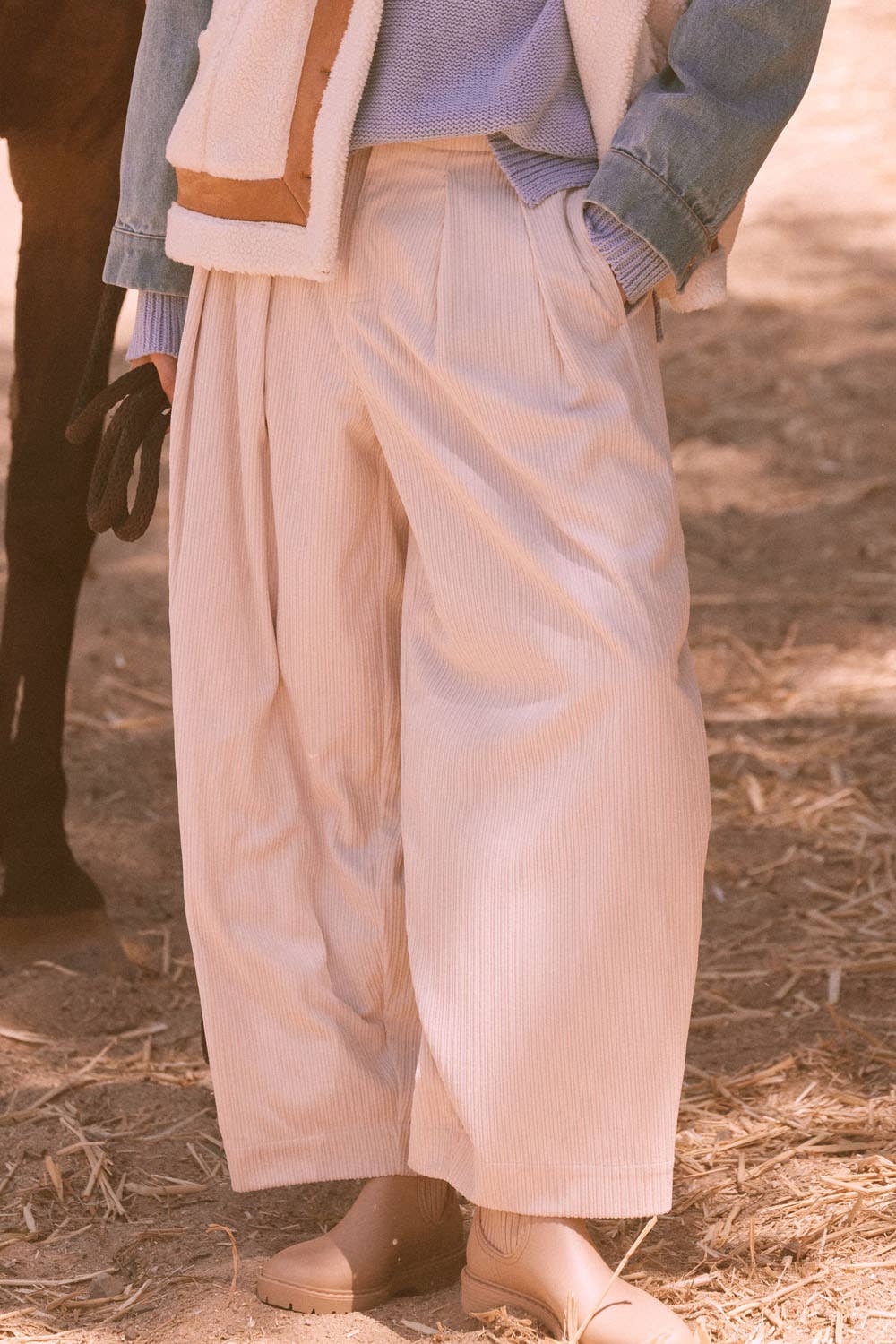 Wearing Promesa USA Velvet Corduroy Pleat-Front Wide-Leg Pants, beige ankle boots, a lavender sweater, denim jacket, and shearling vest, a person stands outdoors on dirt ground with one hand in their pocket.