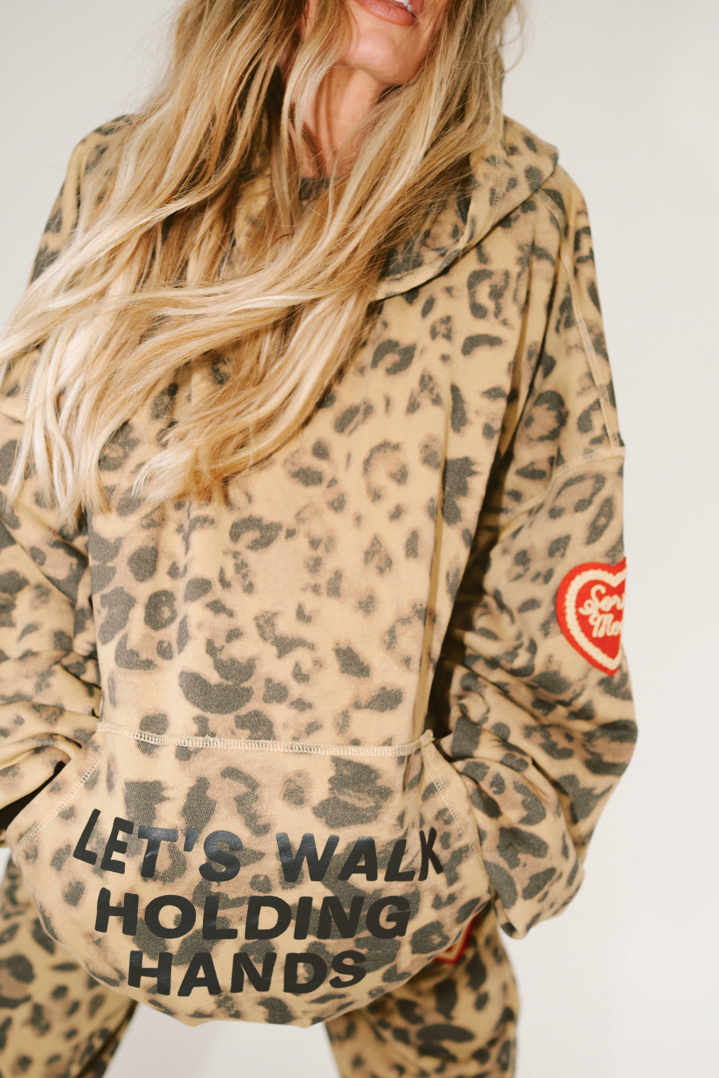 Woman wearing leopard print SORRY MOM HOODIE - PRESALE with text "LET'S WALK HOLDING HANDS" on front pocket and heart patch on sleeve