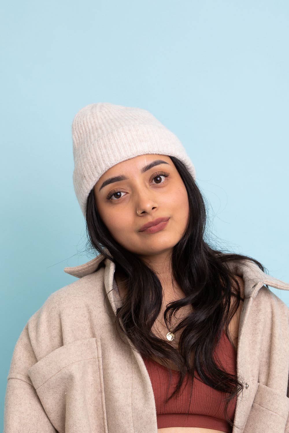 A young woman with long dark hair wears the Versatile Cuffed Beanie – Cozy Rib Knit Hat by Leto Accessories, along with a beige jacket and rust-colored top, standing against a light blue background with a neutral expression.